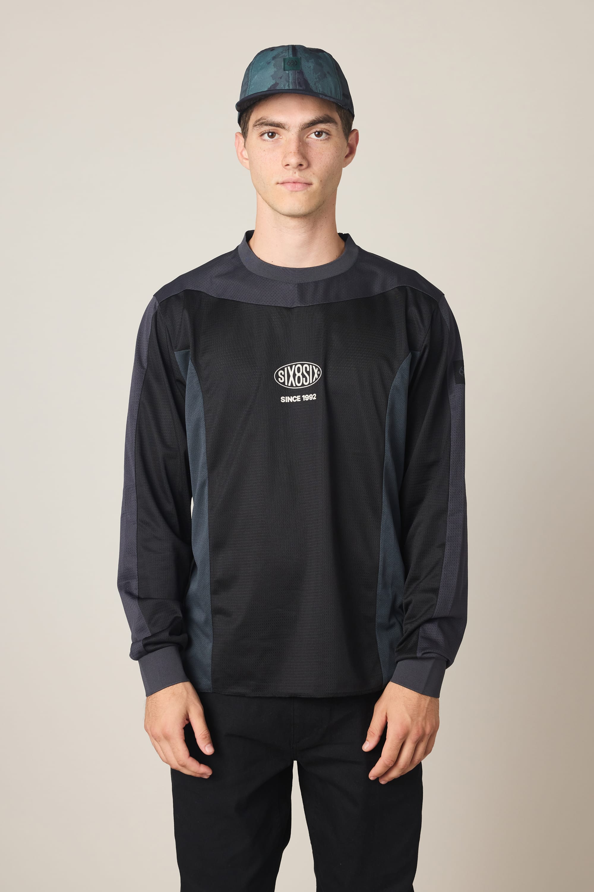 A young man in a teal and black hat and the 686 Men's 92 Turbo LS Tech Tee, featuring SIXEIGHT and SINCE 1992 on the chest, stands against a plain light background.