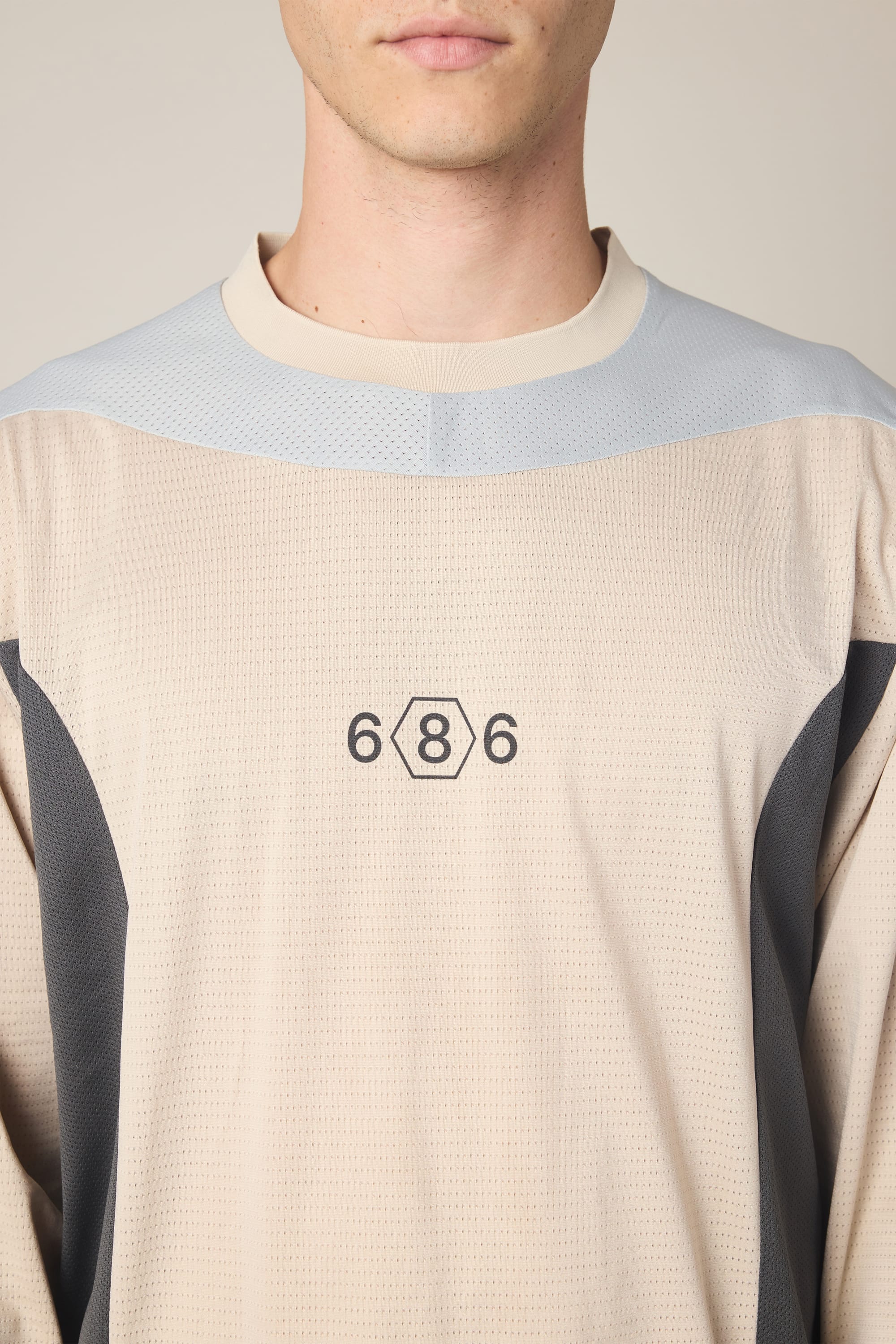 A person is shown from the lower face to upper torso wearing the 686 Men's Hex Turbo LS Tech Tee, a beige and gray polyester shirt featuring a hexagon design with the numbers "686" on the chest.