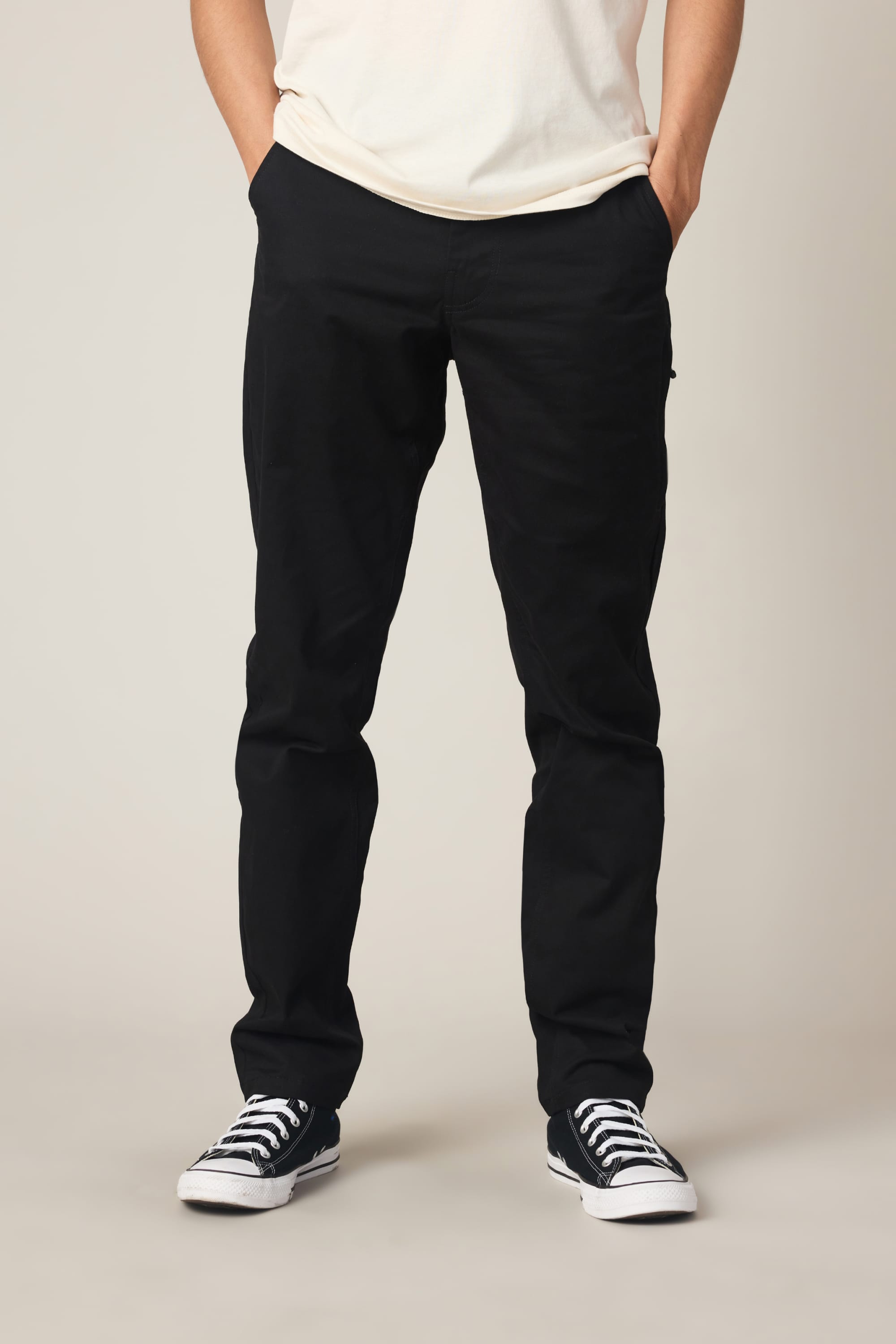 A person stands with hands in pockets against a light background, wearing a white shirt, black-and-white sneakers, and the 686 Men's Everywhere® Standard Pant - Slim Fit by 686.