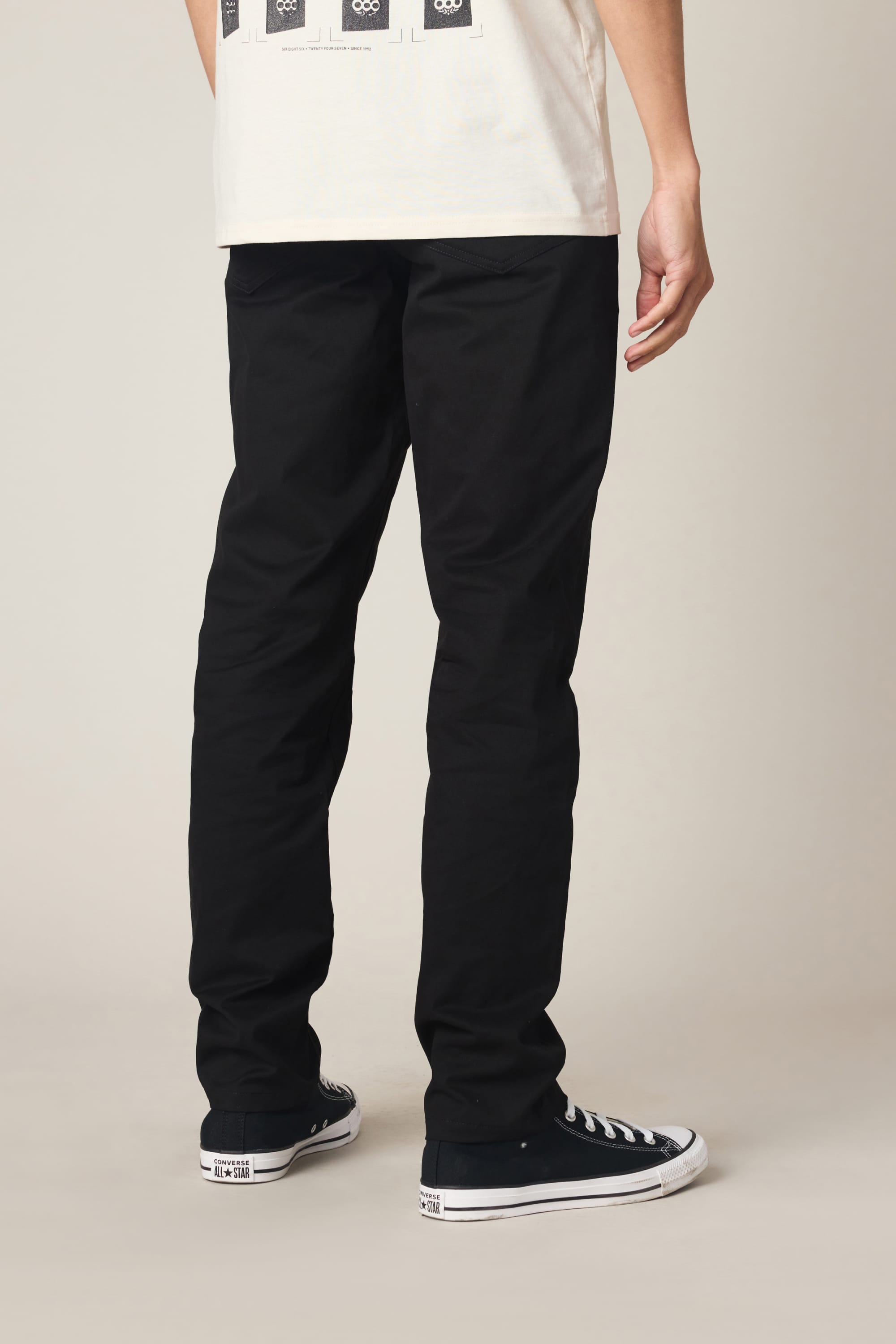 A person faces away, wearing 686 Men's Everywhere® Standard Pant - Slim Fit in black, paired with black low-top Converse sneakers and a light-colored graphic print t-shirt. The setting features a plain, neutral background.