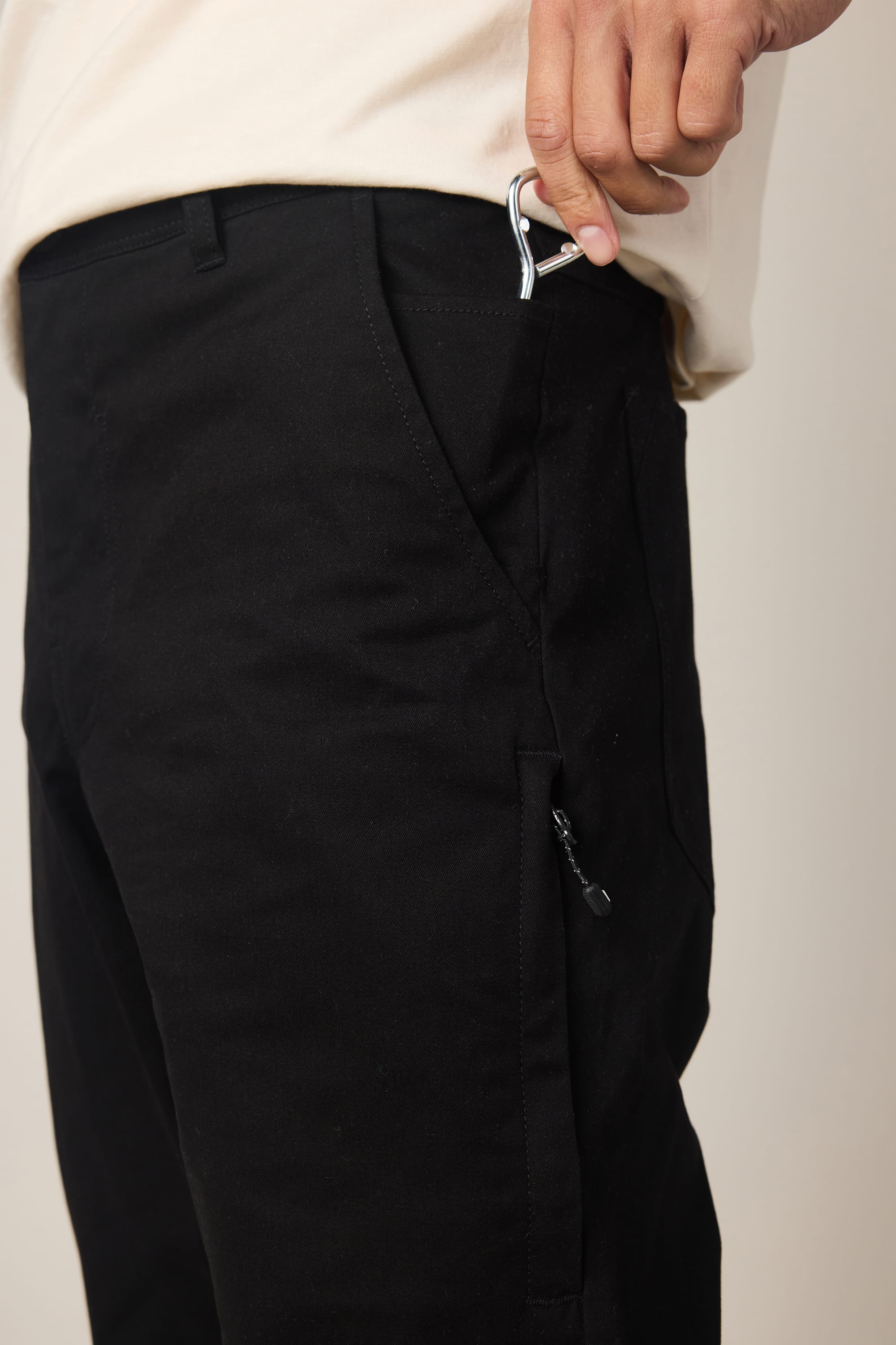 A person wearing 686 Men's Everywhere® Standard Pant - Slim Fit in black and a light shirt puts a small silver carabiner into the front pocket of their pants.