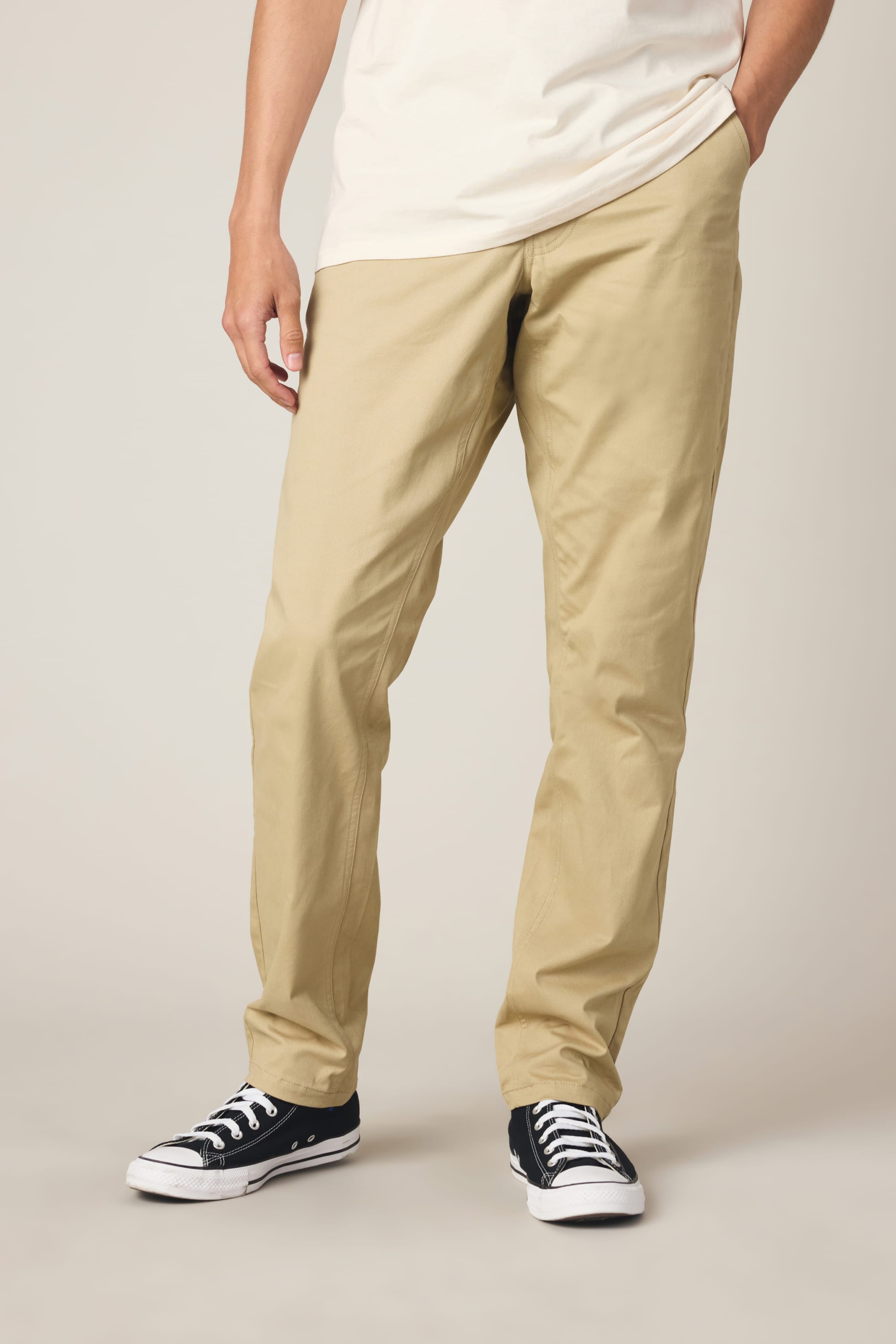 A person wears 686 Men's Everywhere® Standard Pant - Slim Fit in beige, a white T-shirt, and black low-top sneakers, standing with one hand in their pocket against a plain light background. The face is not visible.