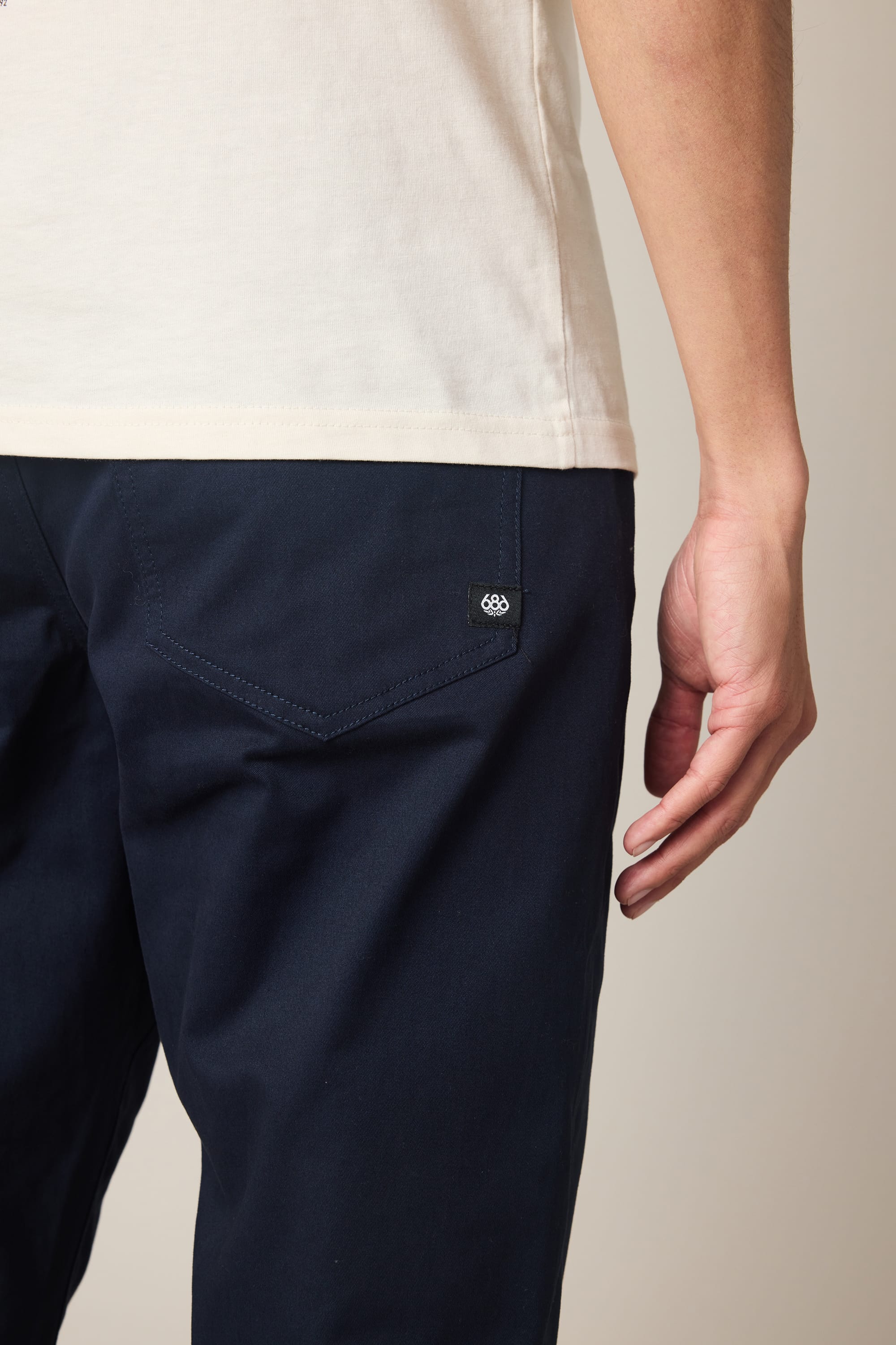 A person wearing a white shirt and the 686 Men's Everywhere® Standard Pant - Slim Fit in dark blue stands sideways, with their right arm relaxed at their side. The image highlights the pants' back pocket and part of the hand.