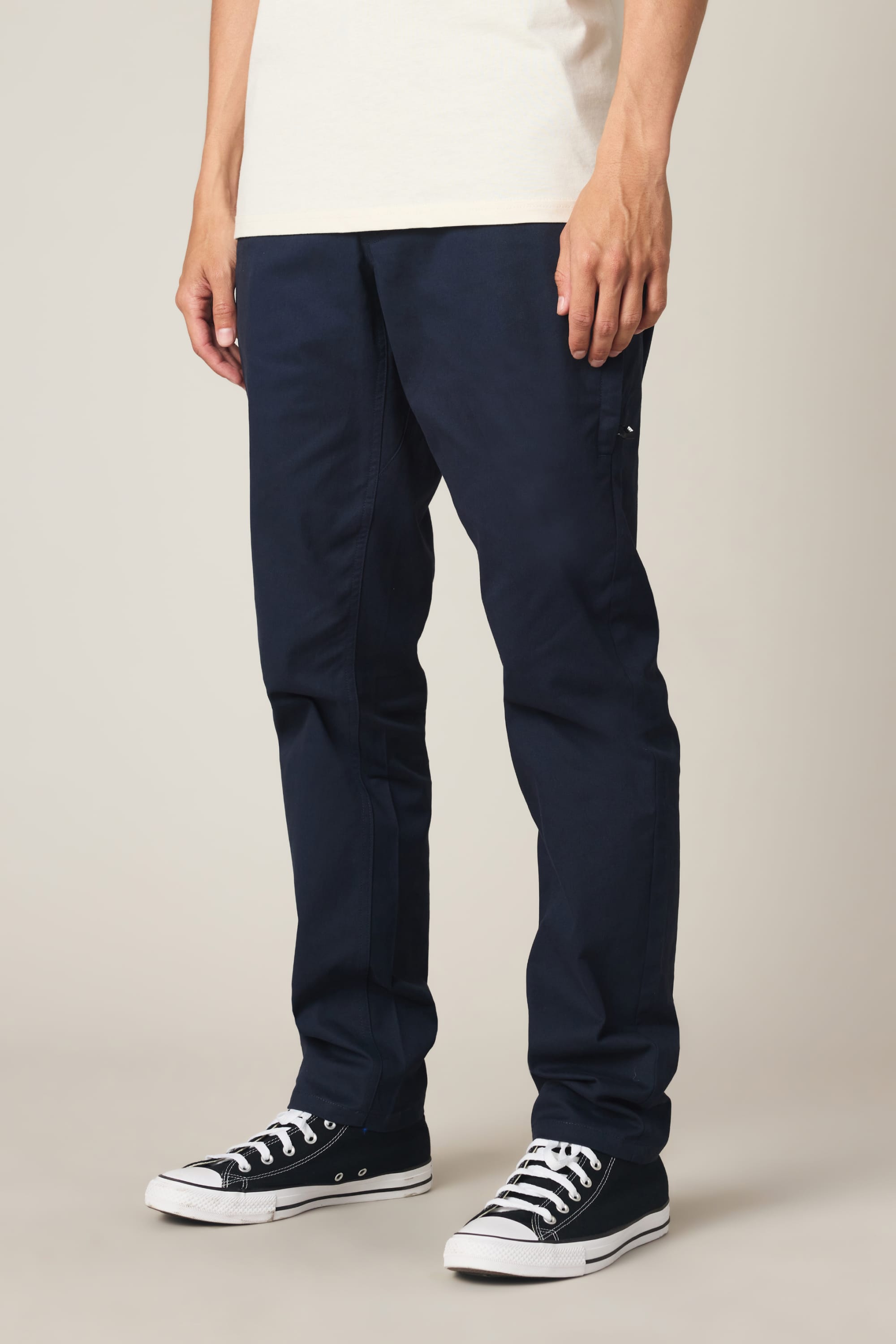 A person is standing against a plain light background, with only the lower half of their body visible. They are wearing a white shirt, black low-top Converse sneakers, and the 686 Men's Everywhere® Standard Pant - Slim Fit from 686 in navy blue, which are crafted from comfortable nylon woven fabric.