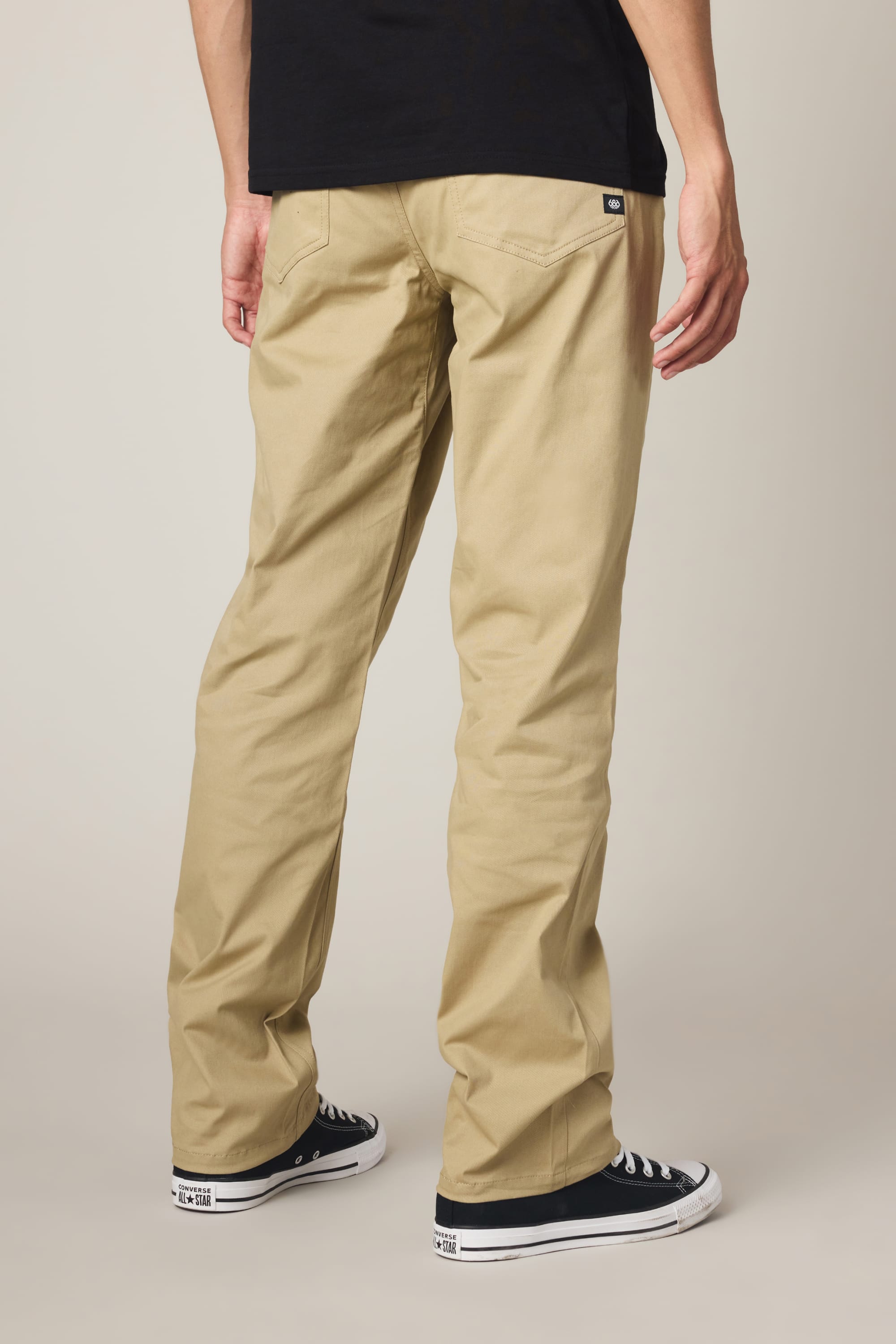 A person stands facing away, wearing a black shirt, black low-top sneakers with white soles, and 686 Men's Everywhere® Standard Pant - Relaxed Fit by 686 in beige with a khaki finish, against a plain light background.