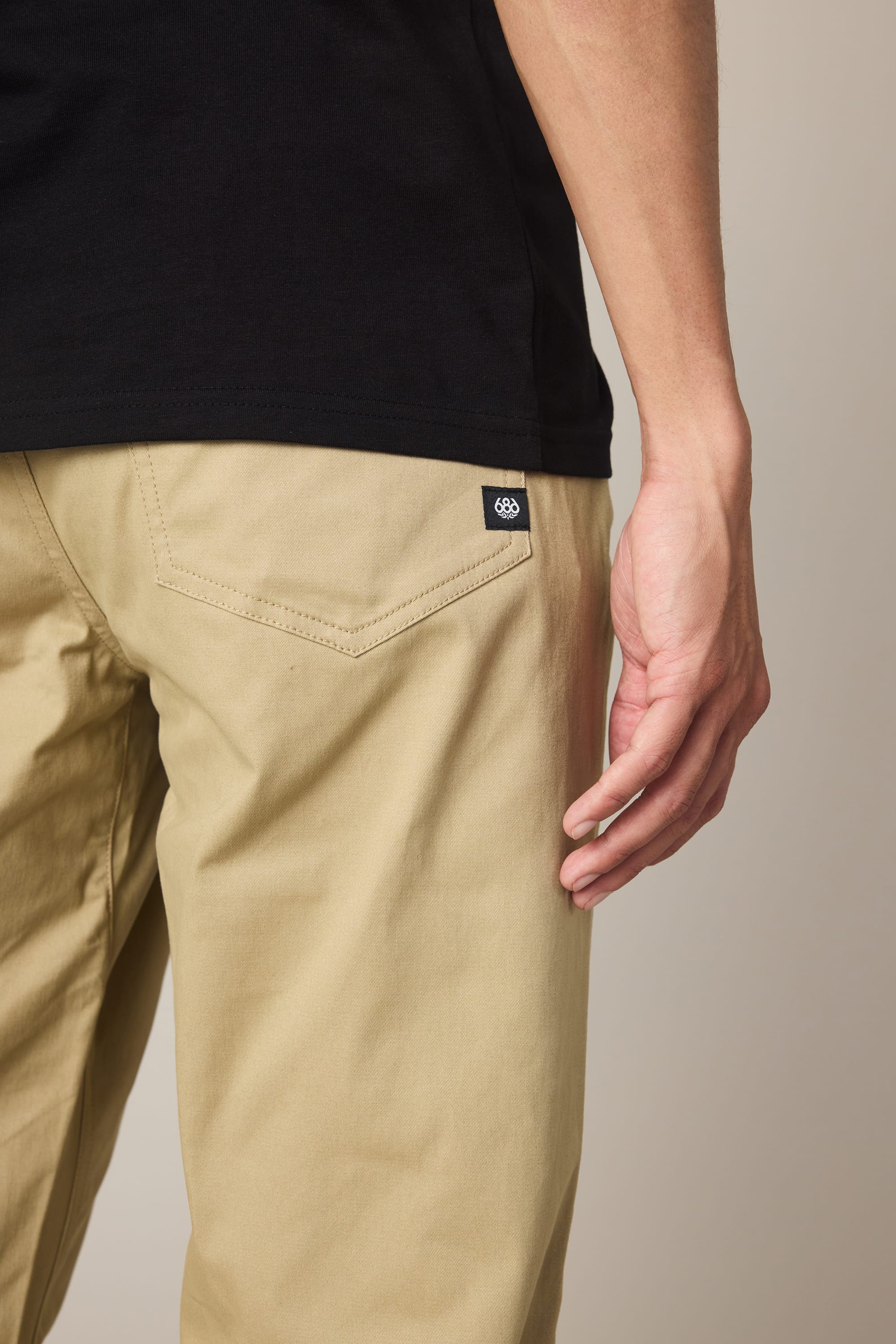 A person wearing 686 Men's Everywhere® Standard Pant - Relaxed Fit in tan and a black shirt stands with their hand by their side. The pant's back pocket features a small black tag with a white circular logo.