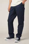 A person wearing 686 Men's Everywhere® Standard Pant - Relaxed Fit in dark blue, a cream t-shirt, and black low-top sneakers stands with one hand in their pocket against a neutral background.