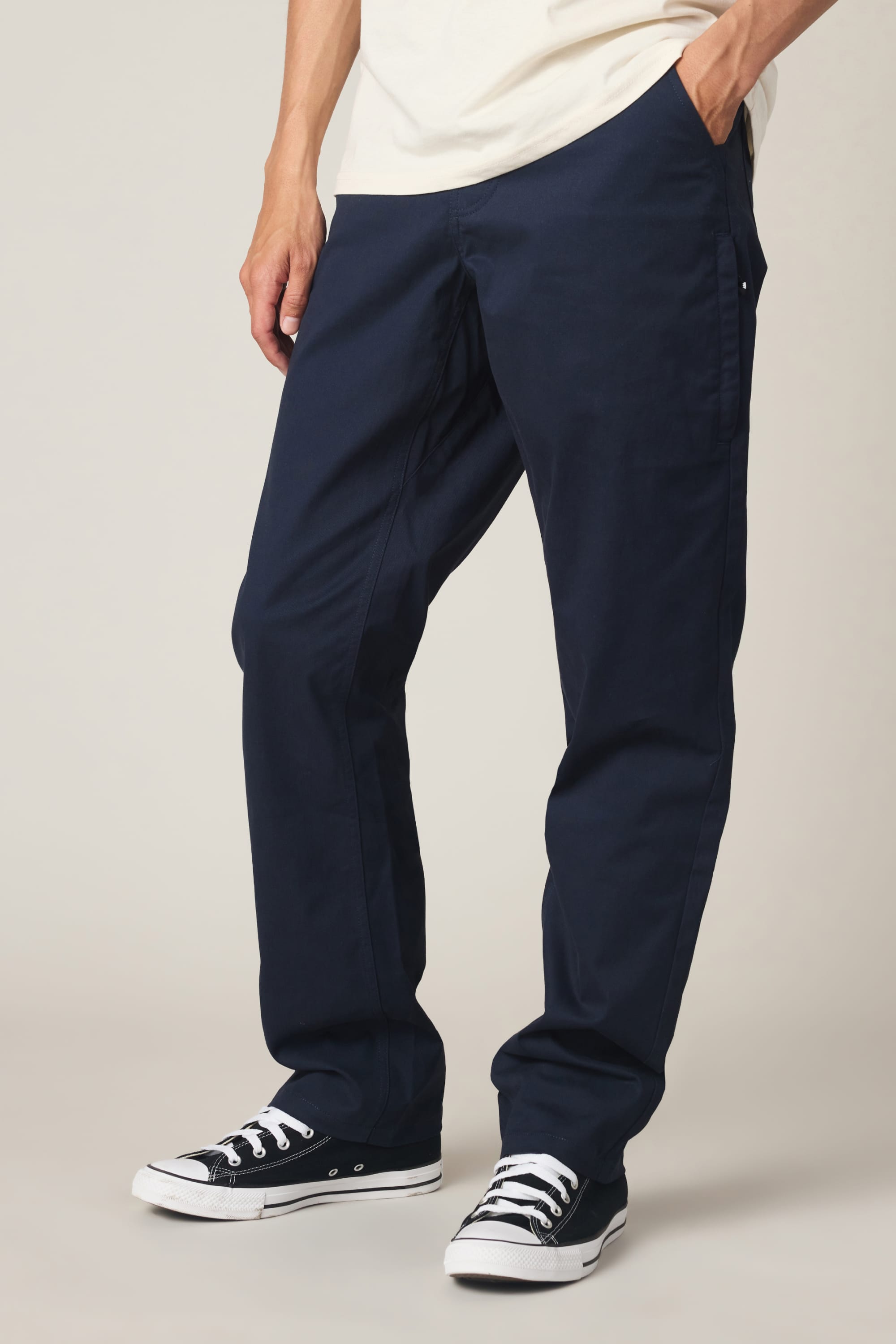 A man wears the 686 Men's Everywhere® Standard Pant - Relaxed Fit in navy blue, paired with black-and-white sneakers, and stands with one hand in his pocket against a plain background.