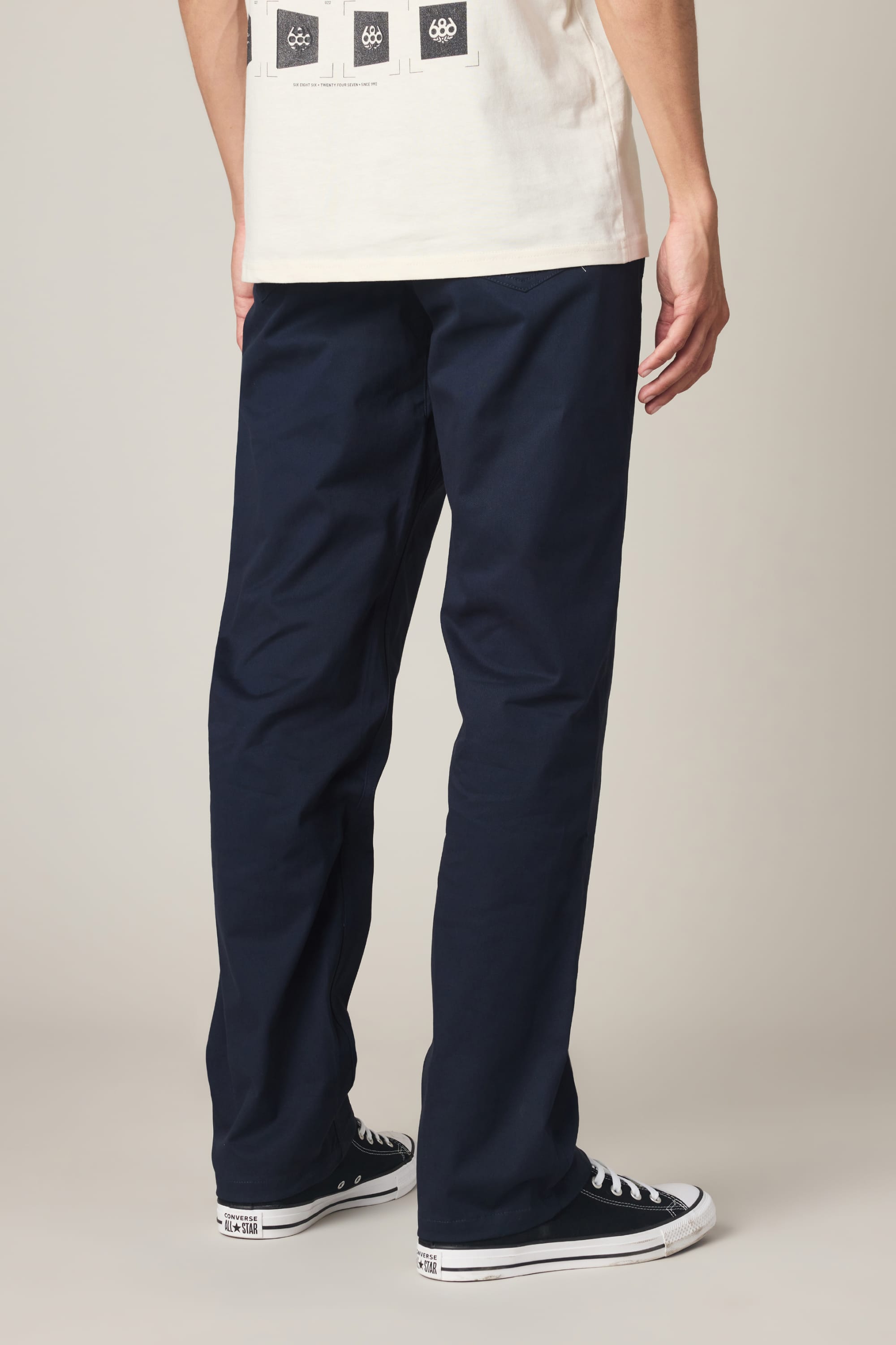 A person is seen from behind wearing the 686 Men's Everywhere® Standard Pant - Relaxed Fit in navy blue by 686, paired with black Converse sneakers. The image shows only the lower torso and legs, with a white shirt partially visible at the top, set against a plain neutral background.
