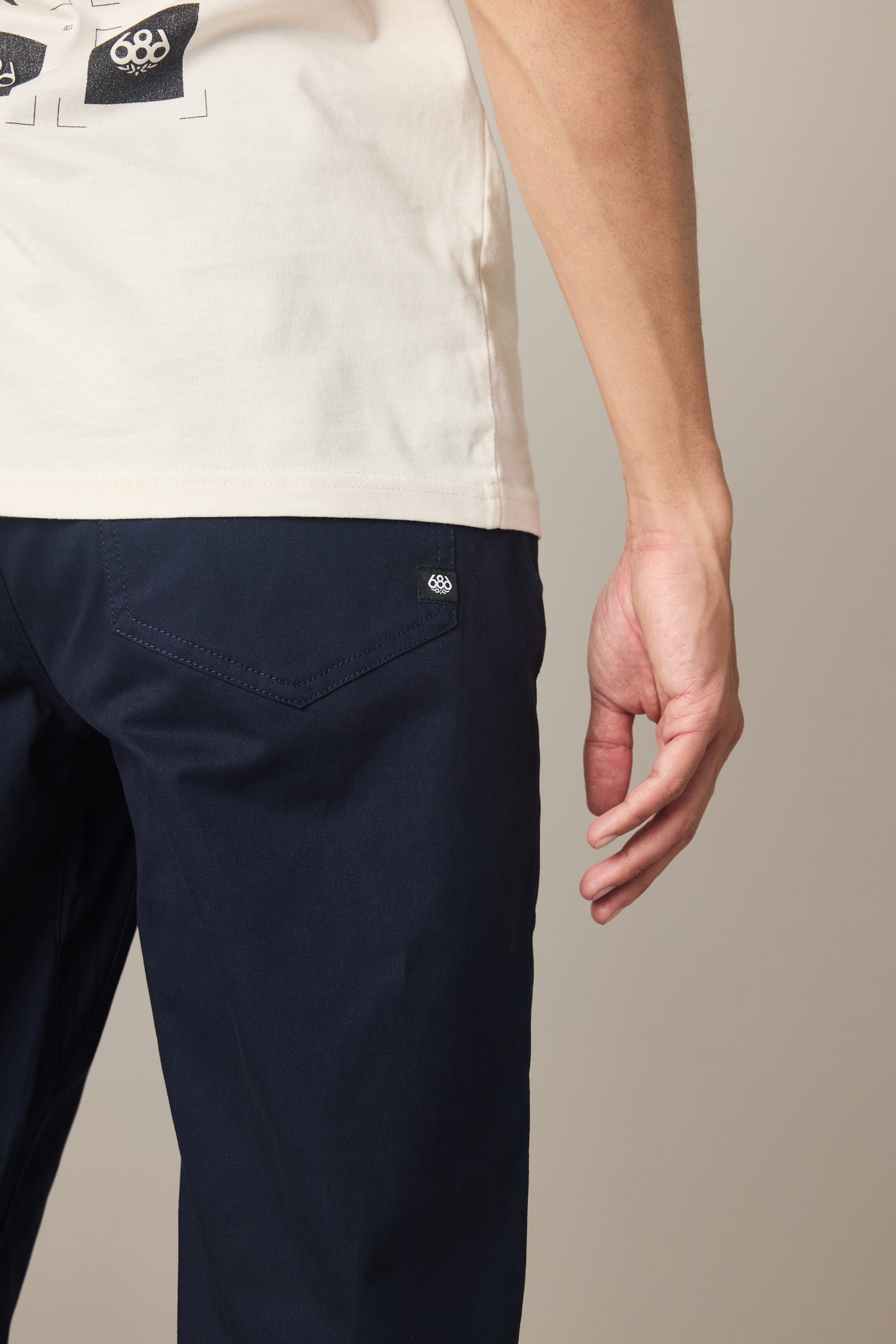 A person wearing a white t-shirt and 686 Men’s Everywhere® Standard Pant - Relaxed Fit (navy blue) stands with their right arm relaxed, highlighting the hip, hand, back pocket, and a small 686 logo on the pants.