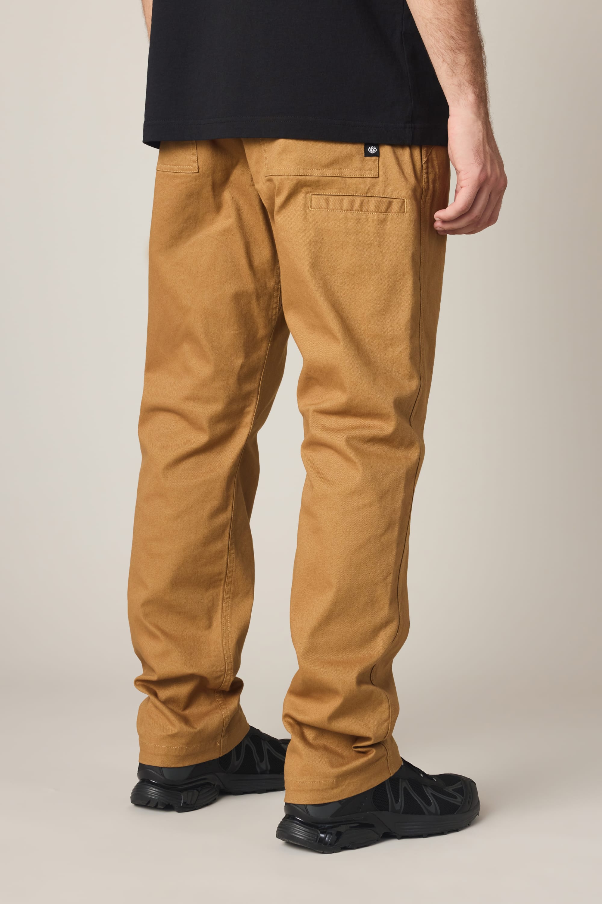 A person wearing 686 Men's Unwork® Tradesman Pant - Relaxed Fit in caramel brown, a black t-shirt, and black shoes stands with their back to the camera. Only the lower half of their body is shown against a plain background.