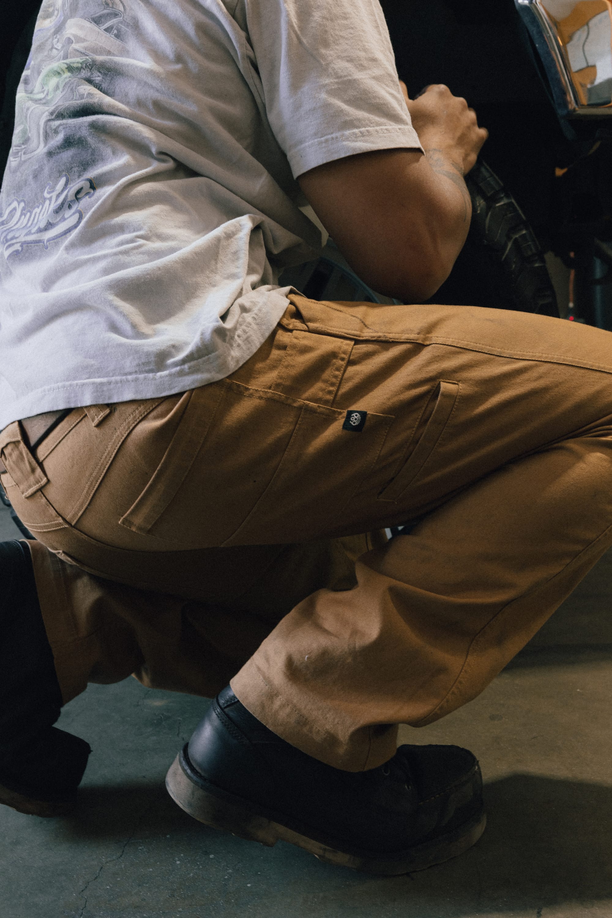 A person wearing the 686 Men's Unwork® Tradesman Pant - Relaxed Fit in brown and black boots is crouching on a concrete floor, holding a tire. Only the lower half of their body is visible.