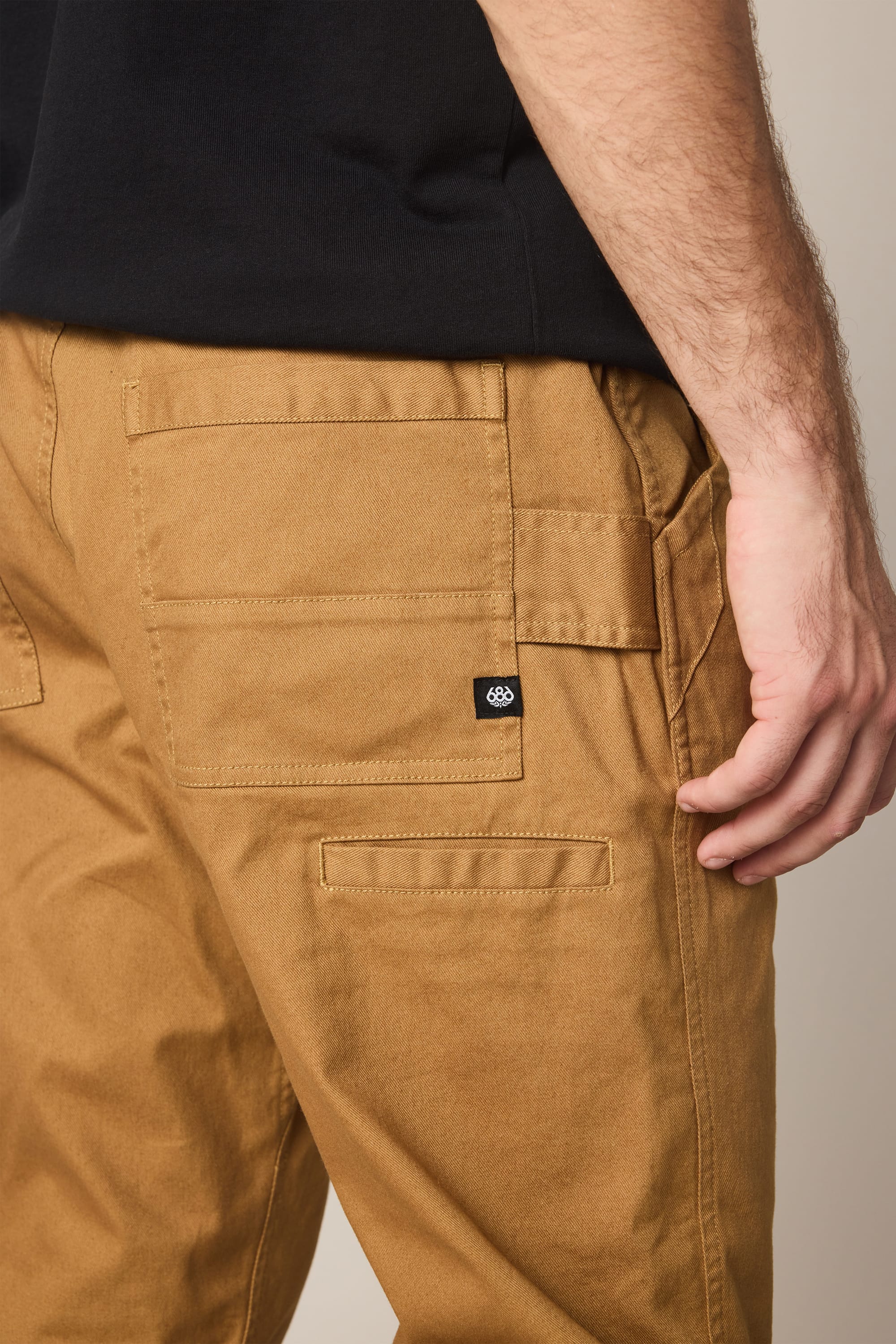 A close-up of a person wearing 686 Men's Unwork® Tradesman Pant - Relaxed Fit in tan by 686, paired with a black shirt, highlighting the side and back pocket details of the pants.