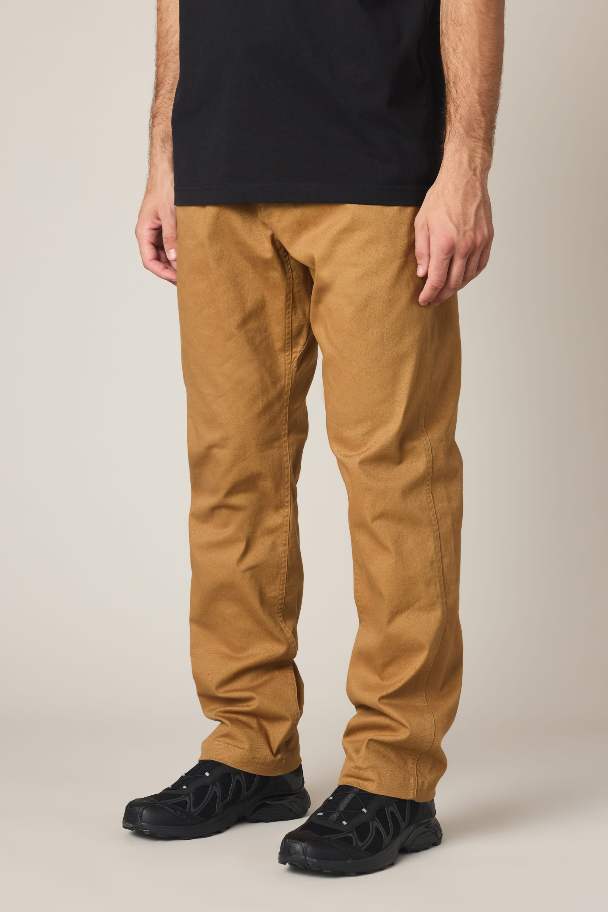 A person is shown from the waist down against a plain background, wearing 686 Men's Unwork® Tradesman Pant - Relaxed Fit in tan, paired with a black t-shirt and black sneakers.