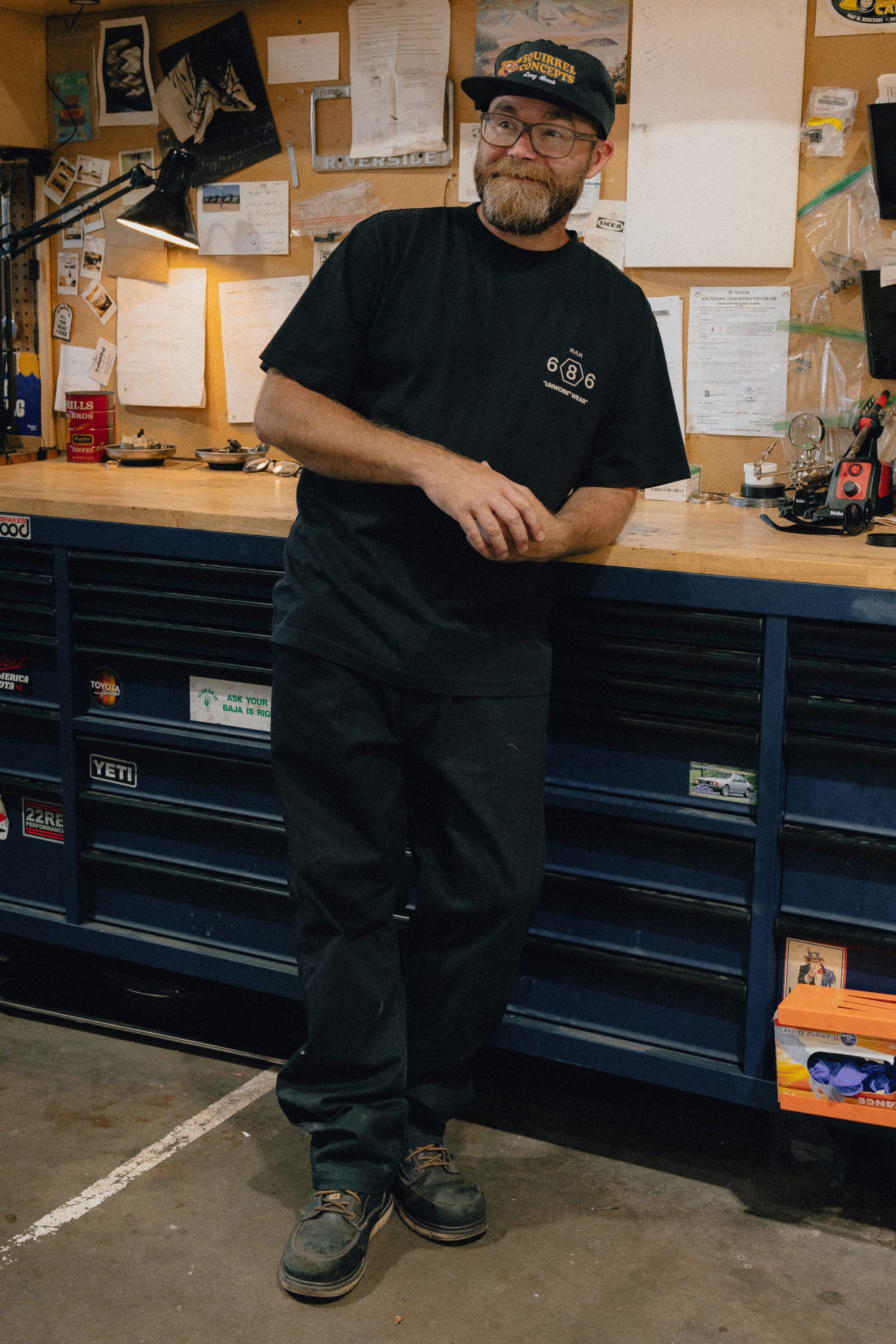 A man with a beard and glasses, wearing a black hat, black T-shirt, and the 686 Men's Unwork® Tradesman Pant - Relaxed Fit by 686, leans casually against a blue tool chest in a workshop with papers and tools on the wall behind him.