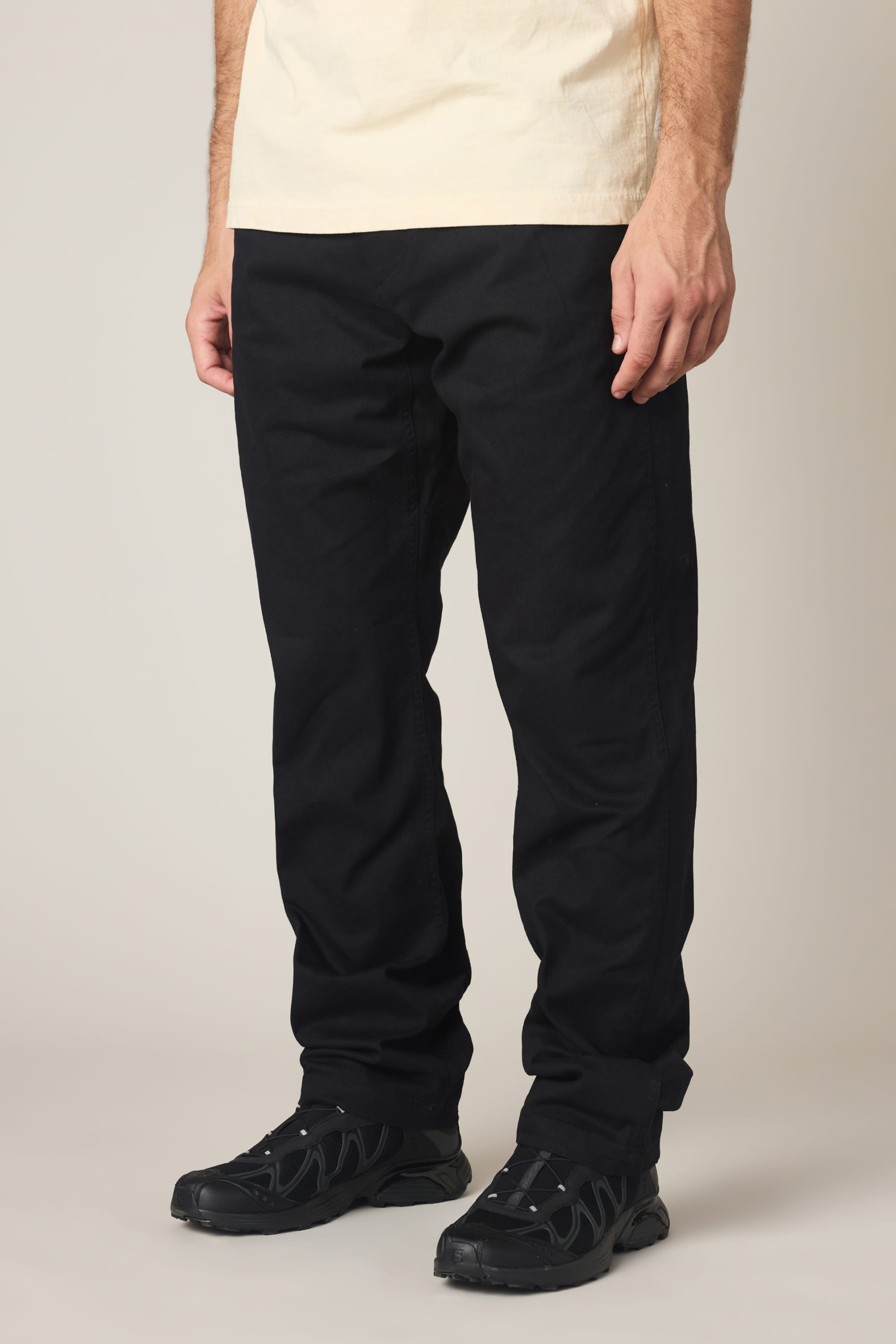 A person wearing a cream shirt, 686 Men's Unwork® Tradesman Pant - Relaxed Fit by 686 in black, and black sneakers stands against a light background. The photo is cropped from shoulders to feet.