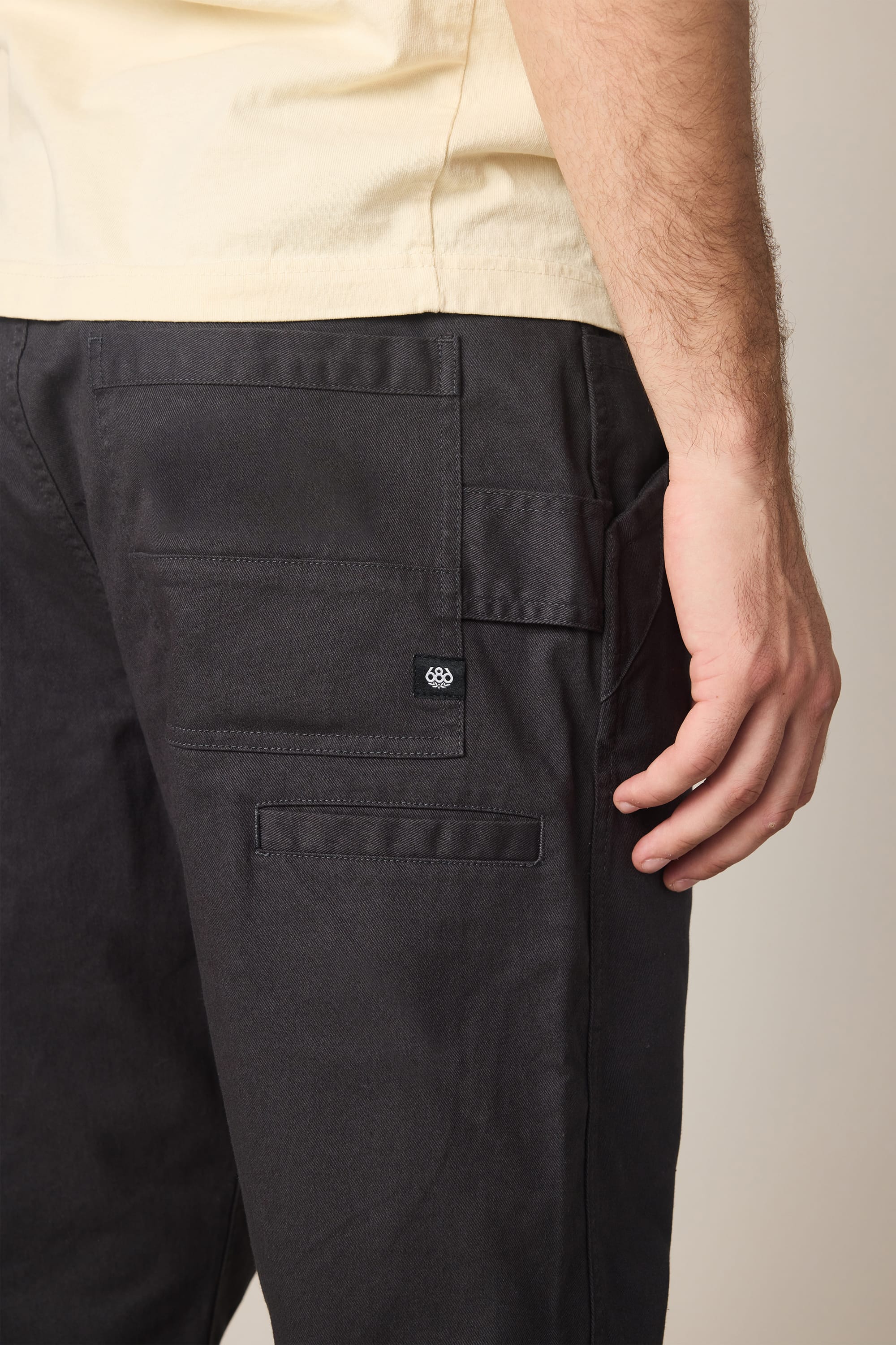 A person is shown from the waist down wearing a light yellow shirt and black 686 Men's Unwork® Tradesman Pant - Relaxed Fit, with their right arm by their side and several pockets visible on the pants.