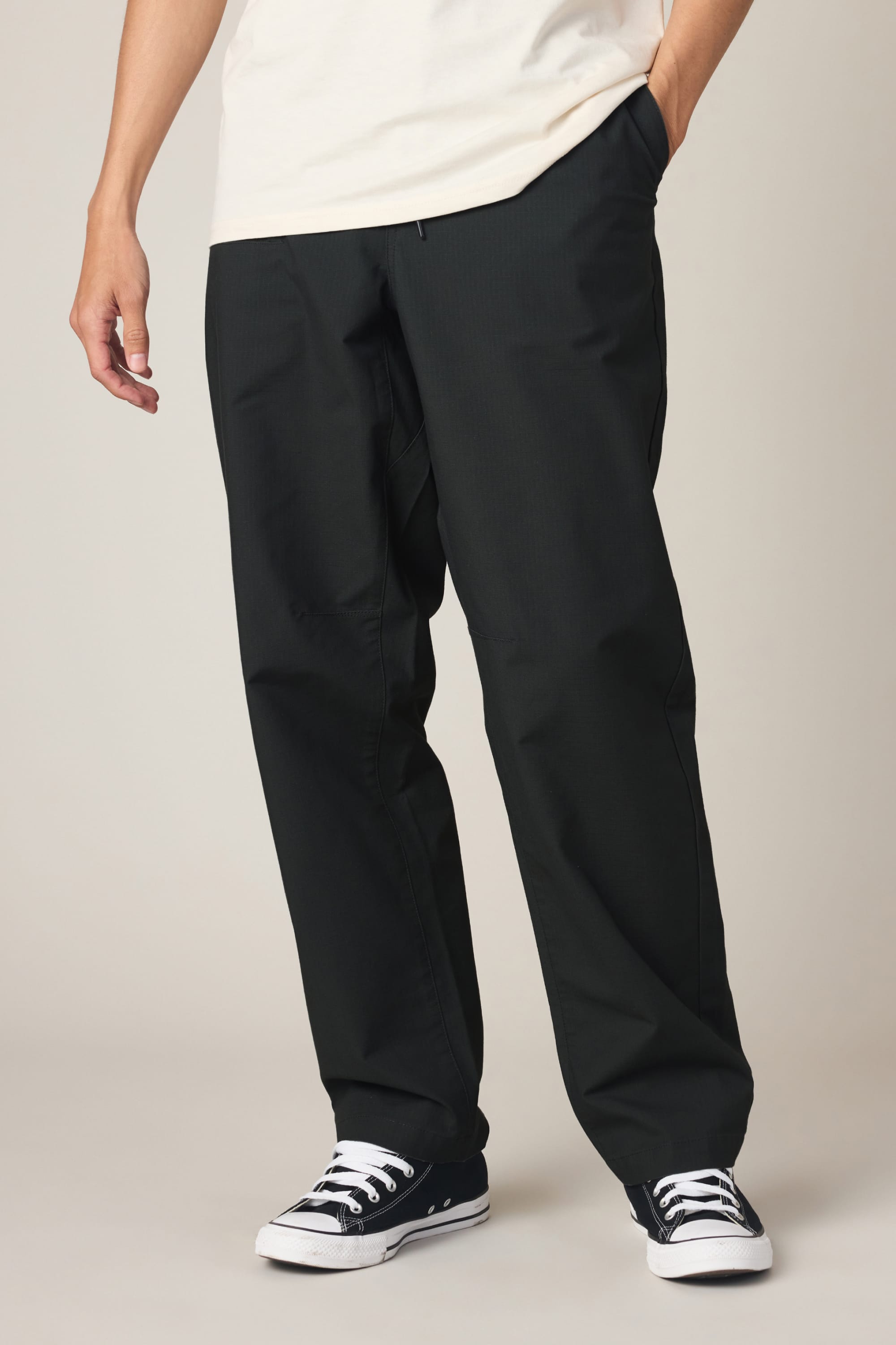 Wearing 686 Men's Free Loader Pant - Loose Fit by 686, a person pairs these pants with a cream shirt and black low-top sneakers with white soles, standing casually with one hand in their pocket against a plain light background.