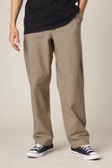 A person wearing 686 Men's Free Loader Pant - Loose Fit in khaki, a black T-shirt, and black low-top sneakers with white soles stands with one hand in their pocket against a neutral background.