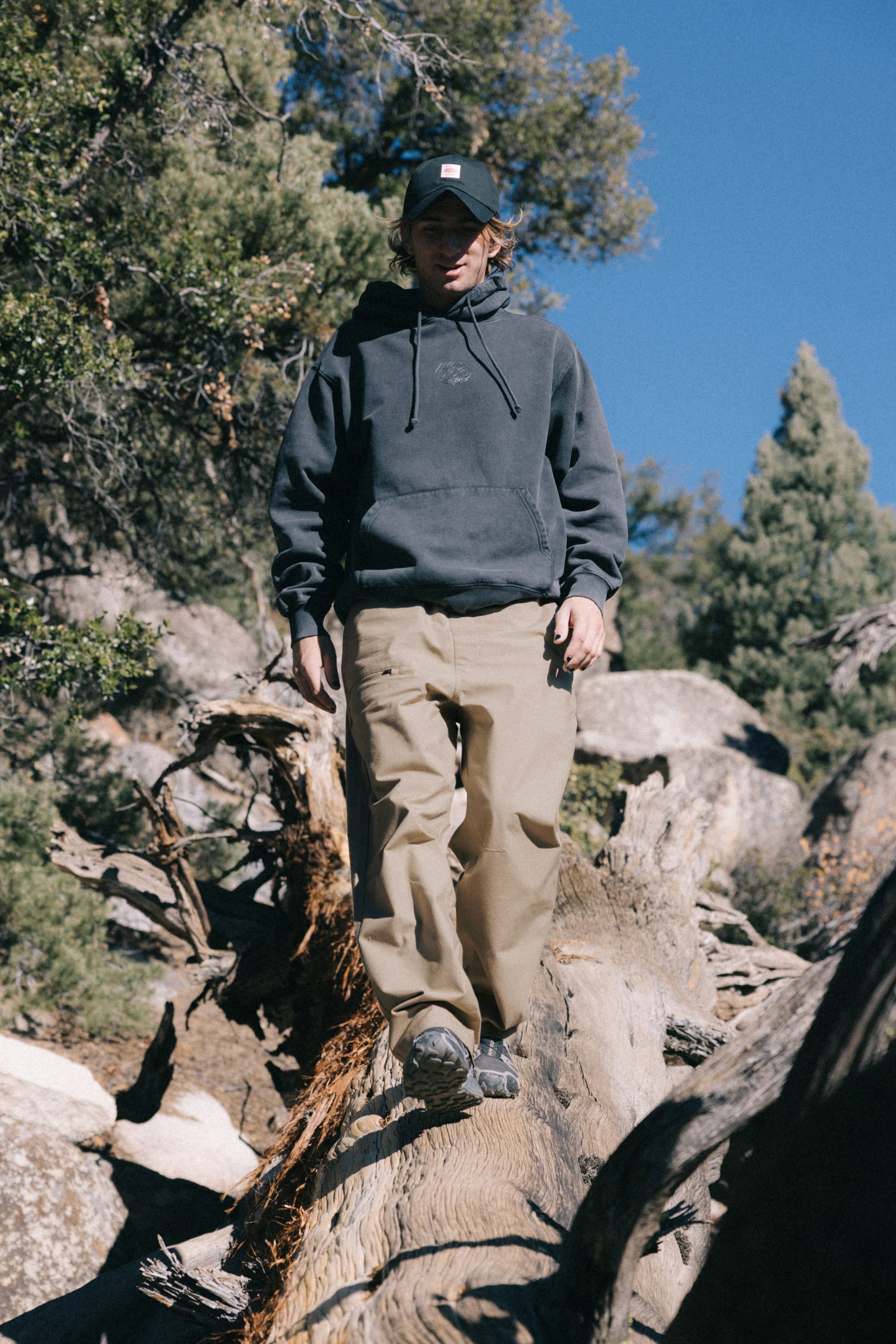A person in a dark hoodie, the 686 Men's Free Loader Pant - Loose Fit by 686, and a cap walks on a fallen tree trunk outdoors, surrounded by rocks and pine trees under a clear blue sky.