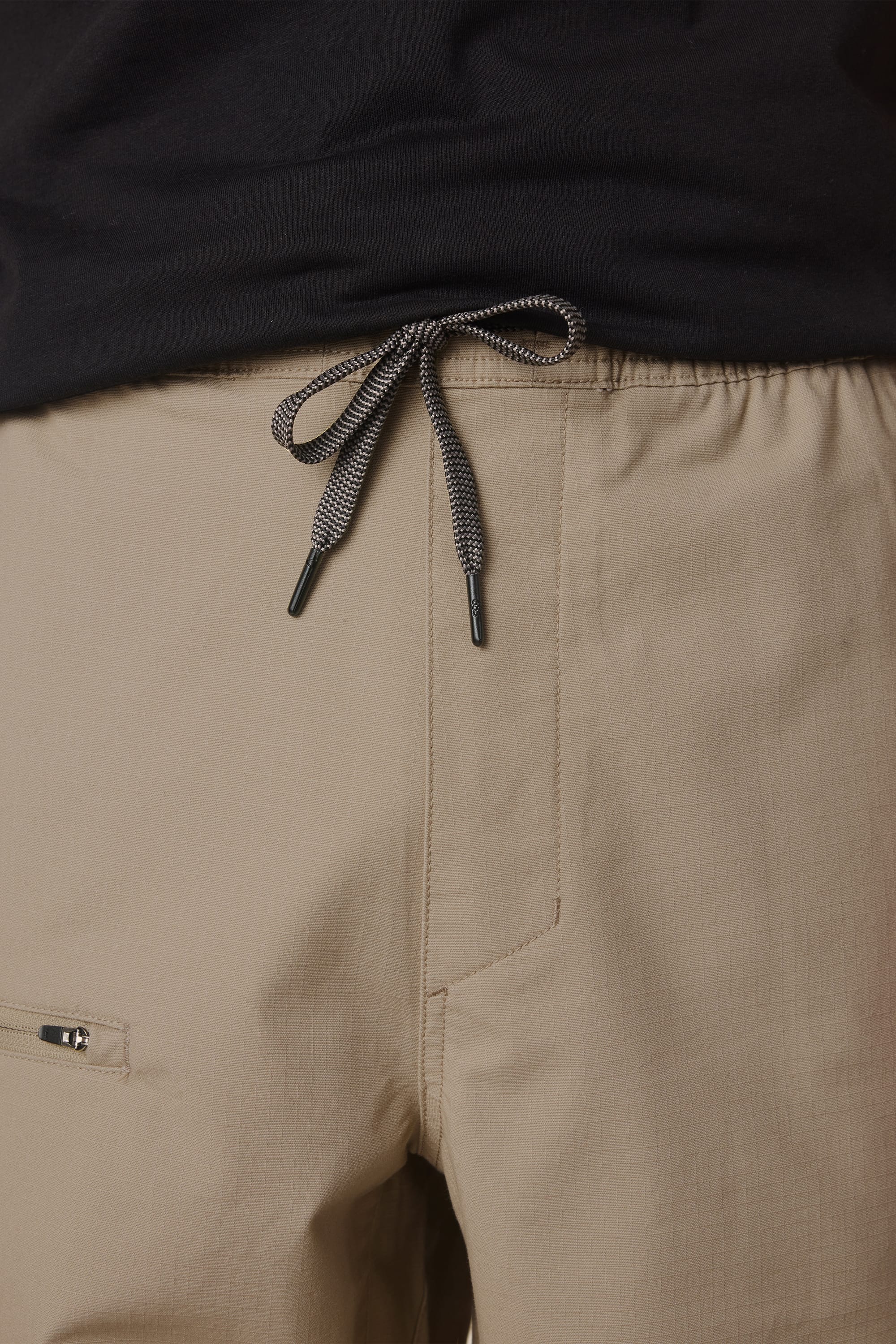 Close-up of a person wearing the 686 Men's Free Loader Pant - Loose Fit in light brown, showing the drawstring waist knot, textured nylon fabric, and part of a zippered pocket—crafted by 686 for durability.