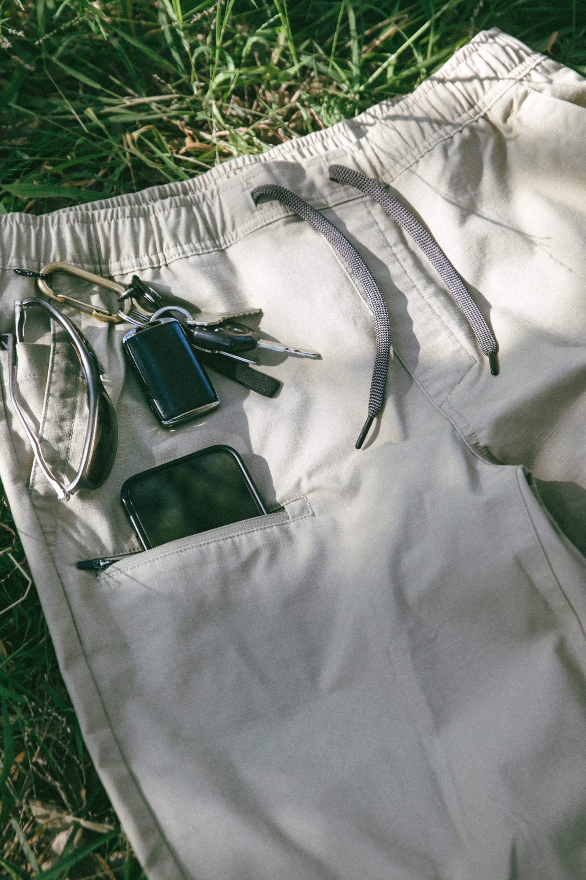 The 686 Men's Free Loader Pant - Loose Fit by 686, featuring khaki spandex woven fabric and a drawstring, is shown laid out on grass. Keys, a black key fob, and a wrist strap are resting on the pants, while a black smartphone is partially visible inside the zipped pocket.