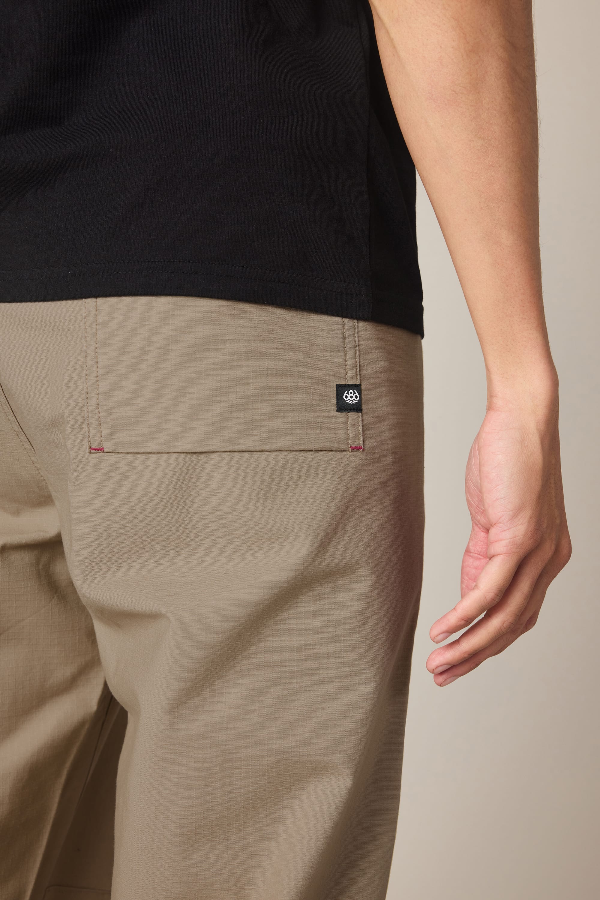 A person wearing a black shirt and the 686 Men's Free Loader Pant - Loose Fit in beige stands with their right arm relaxed, displaying the back pocket and small black and white 686 logo tag.
