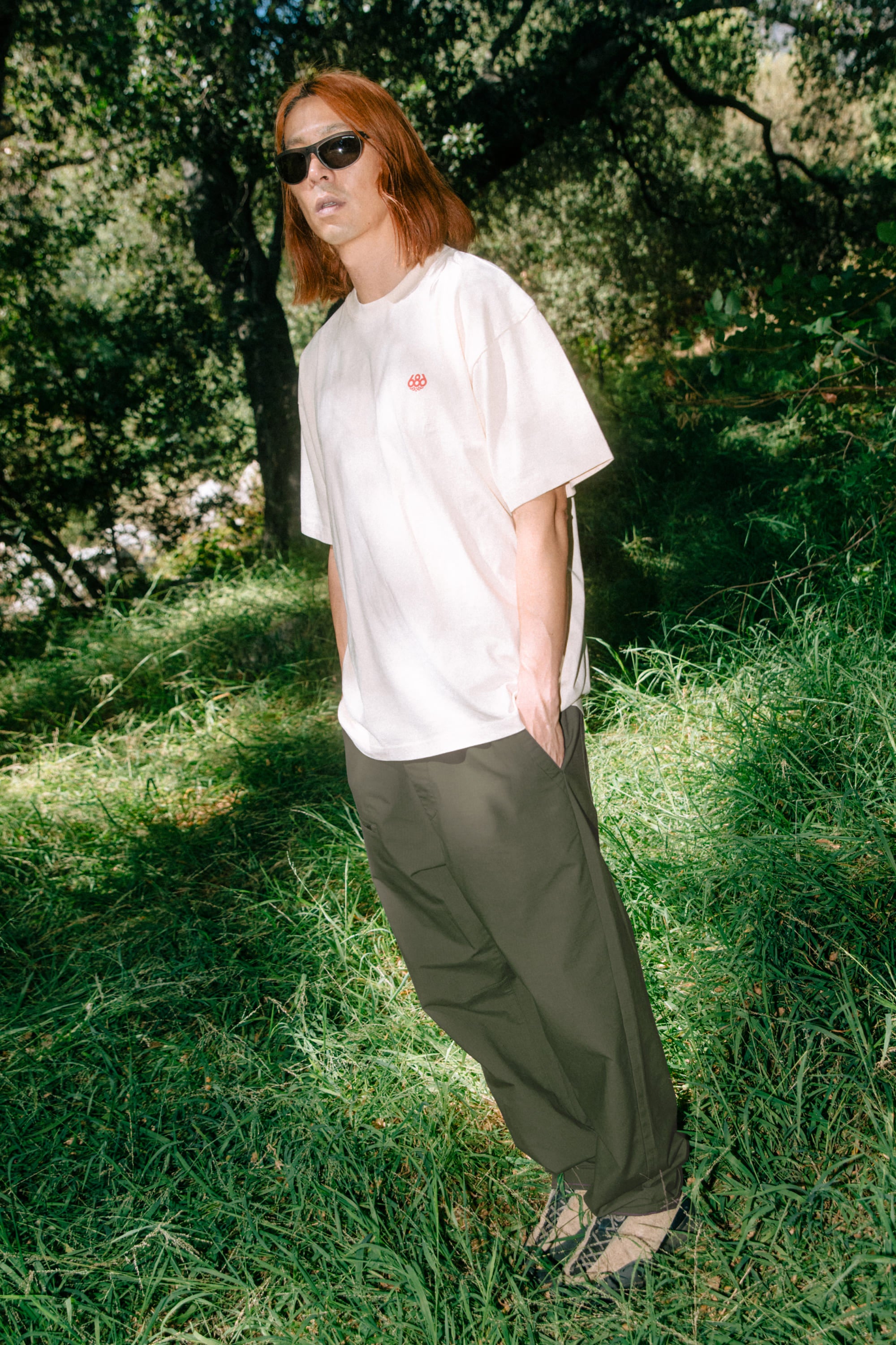 A person with long reddish hair wearing sunglasses, a white T-shirt, and 686 Men's Free Loader Pant - Loose Fit by 686 stands in a grassy, wooded area with hands in their pockets.
