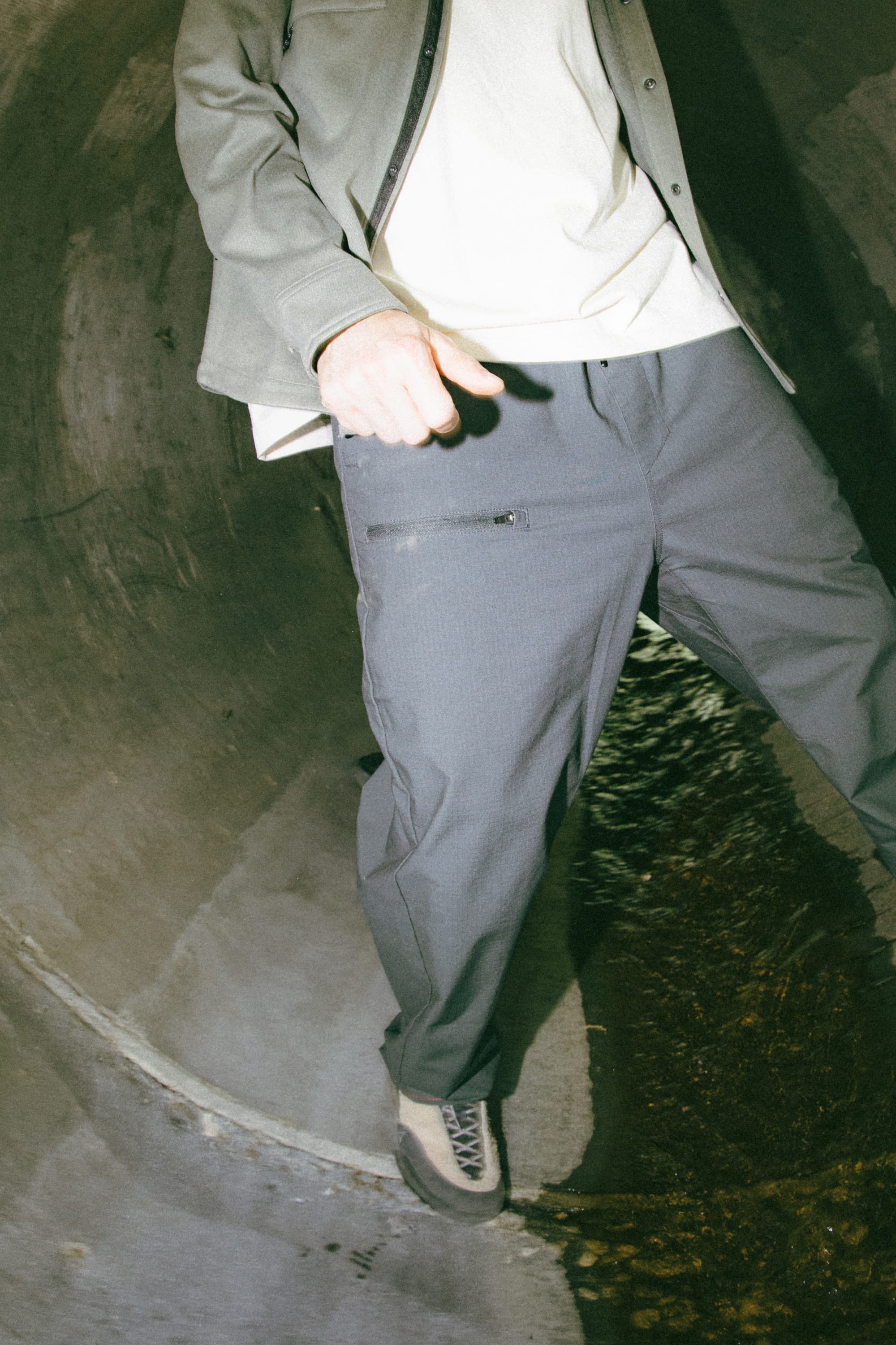 A person wearing a green jacket, white shirt, and the 686 Men's Free Loader Pant - Loose Fit by 686 stands inside a concrete tunnel with water flowing at their feet. Their face is not visible in the image.
