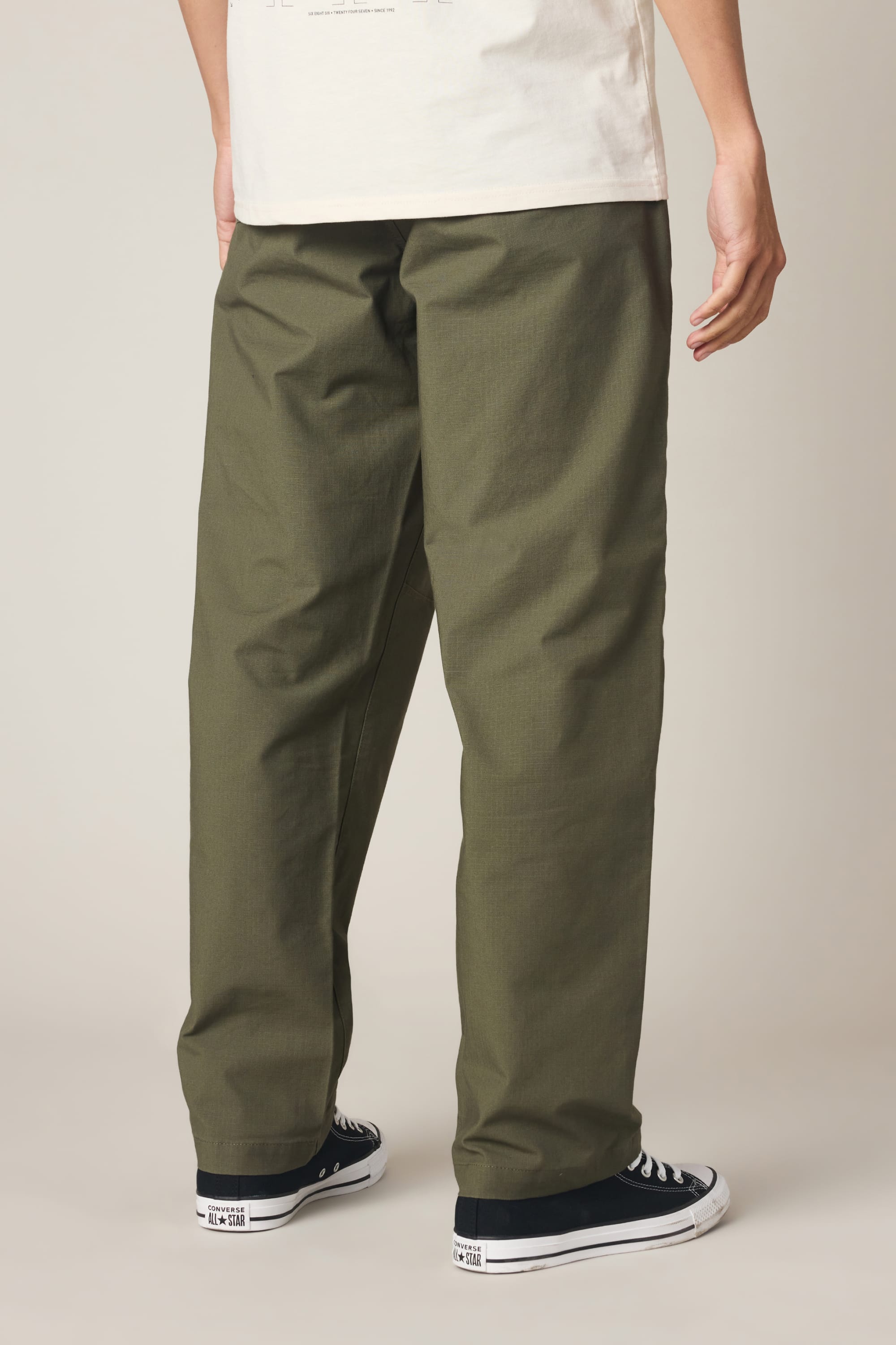 A person stands facing away, wearing 686 Men's Free Loader Pant - Loose Fit in olive green by 686, a white T-shirt, and black low-top sneakers with white soles against a plain light background.
