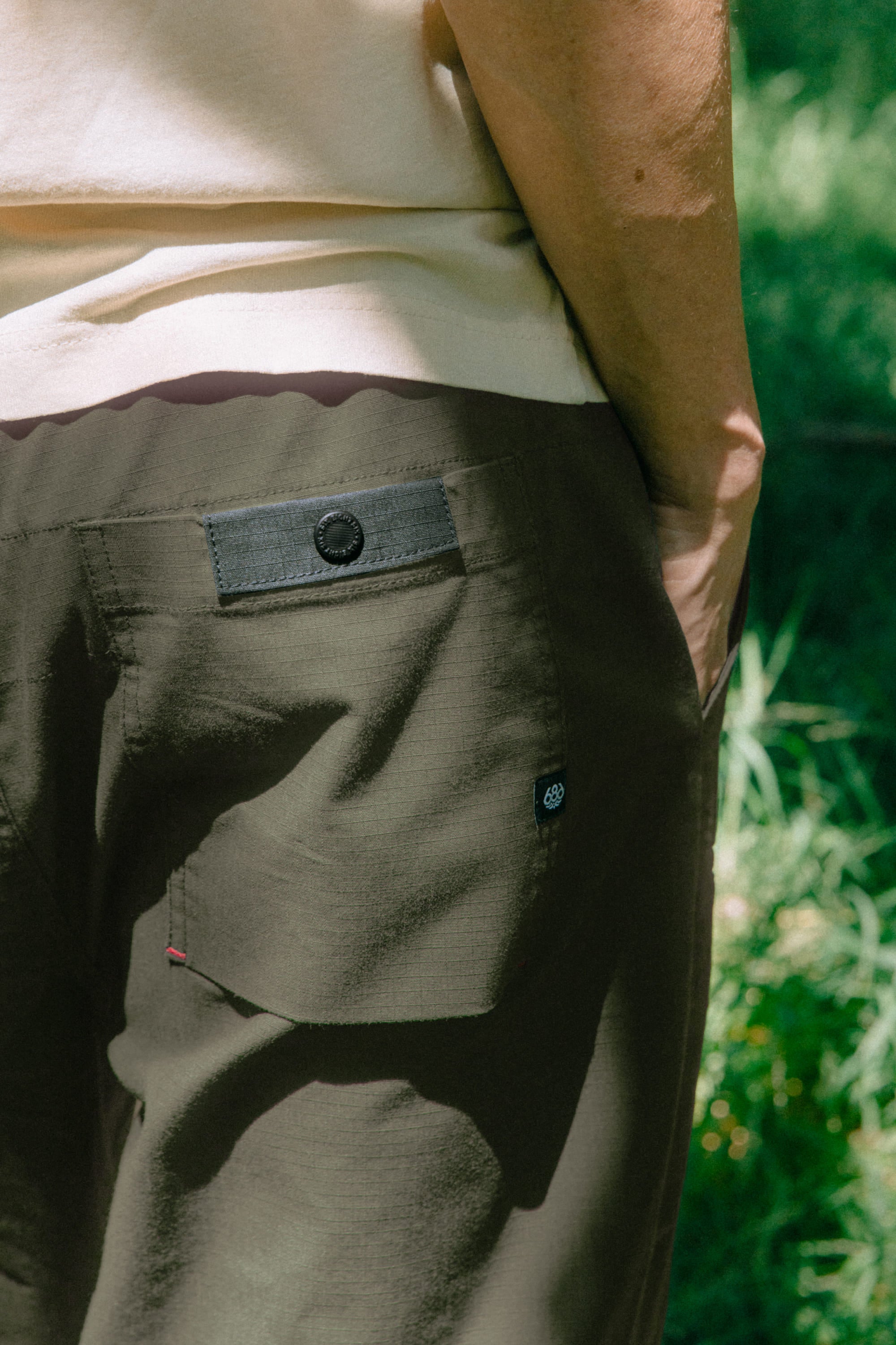 A person stands outdoors in sunlight with greenery in the background, wearing the 686 Men's Free Loader Pant - Loose Fit in olive green, with one hand in the back pocket and part of a beige shirt visible.