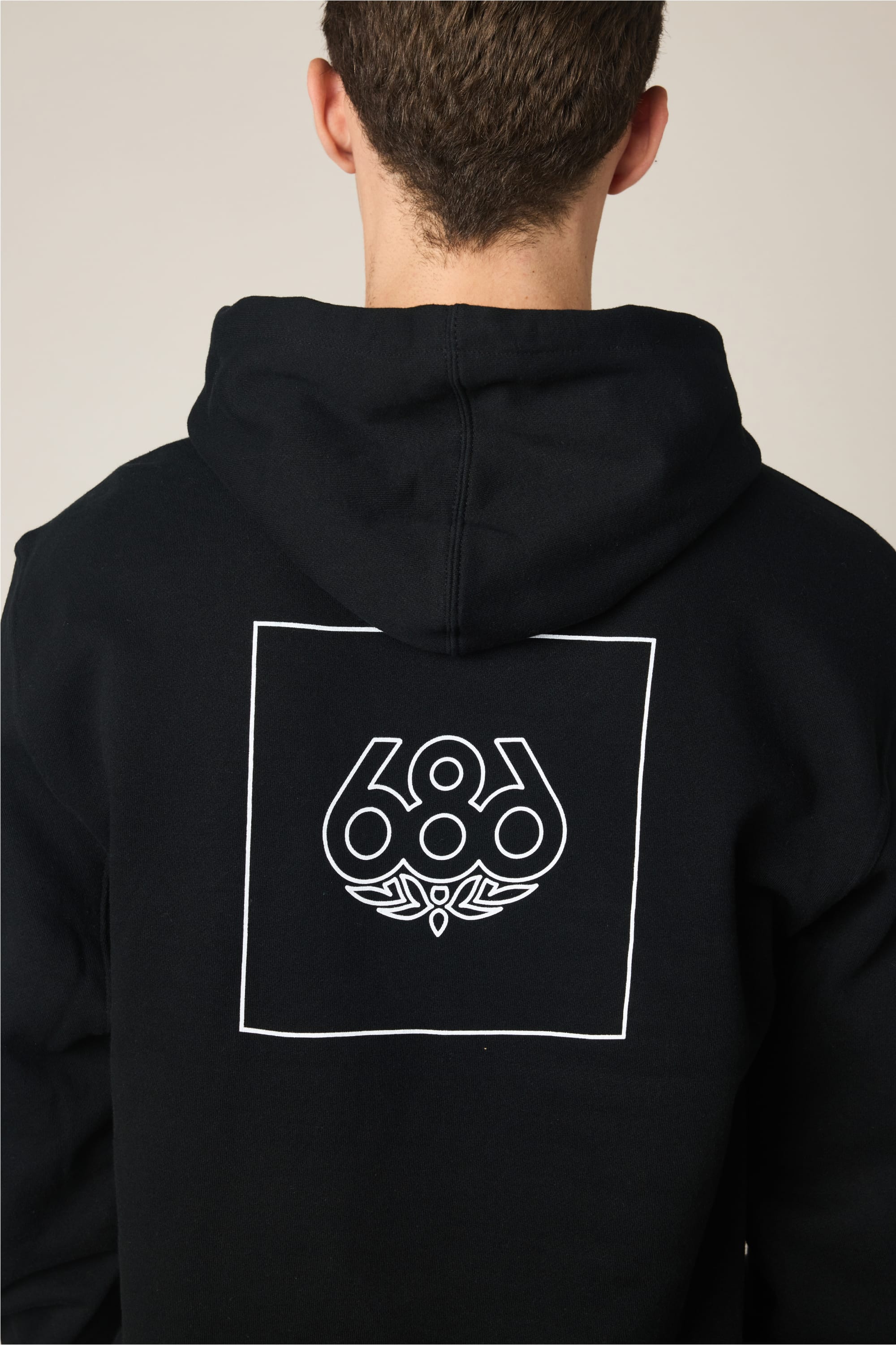 A person seen from behind wears the 686 Wordmark Athletic LS Hoody by 686—a men's cotton sweatshirt with a black hood, featuring a white outlined square and stylized “686” logo on the back.
