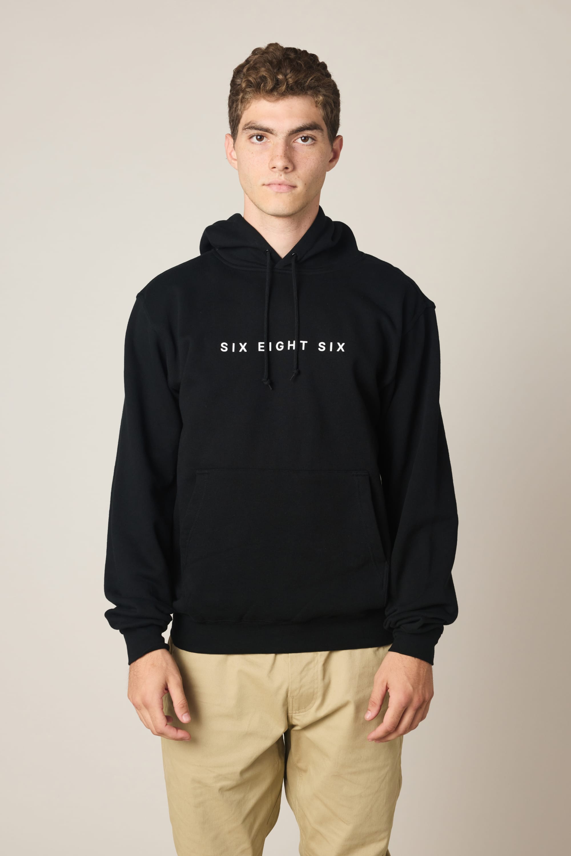 A young person faces forward in a 686 Wordmark Athletic LS Hoody, featuring "686" branding in white on the front, paired with light khaki pants, set against a plain light background.