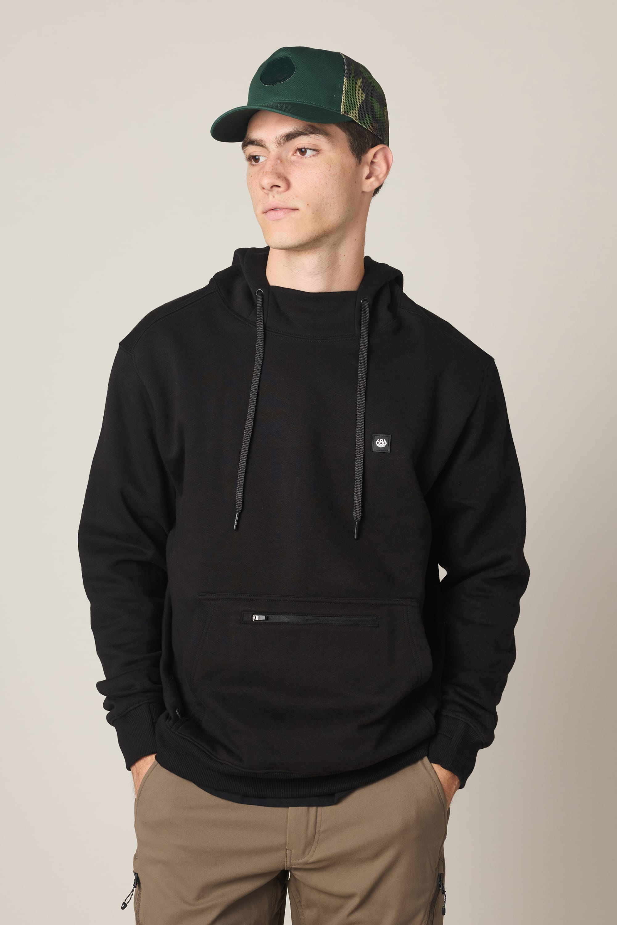 A young man wearing a green camouflage baseball cap, tan pants, and the 686 Men's All Season Hoody by 686 with a small logo stands with his hands in his pockets, looking off to the side against a plain light background.