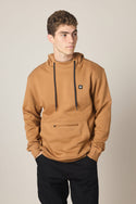 A young man with short curly hair wears the 686 Men's All Season Hoody, featuring black drawstrings and a small black logo patch on the chest, paired with black pants. He stands with hands in the front pocket, looking left against a plain background.