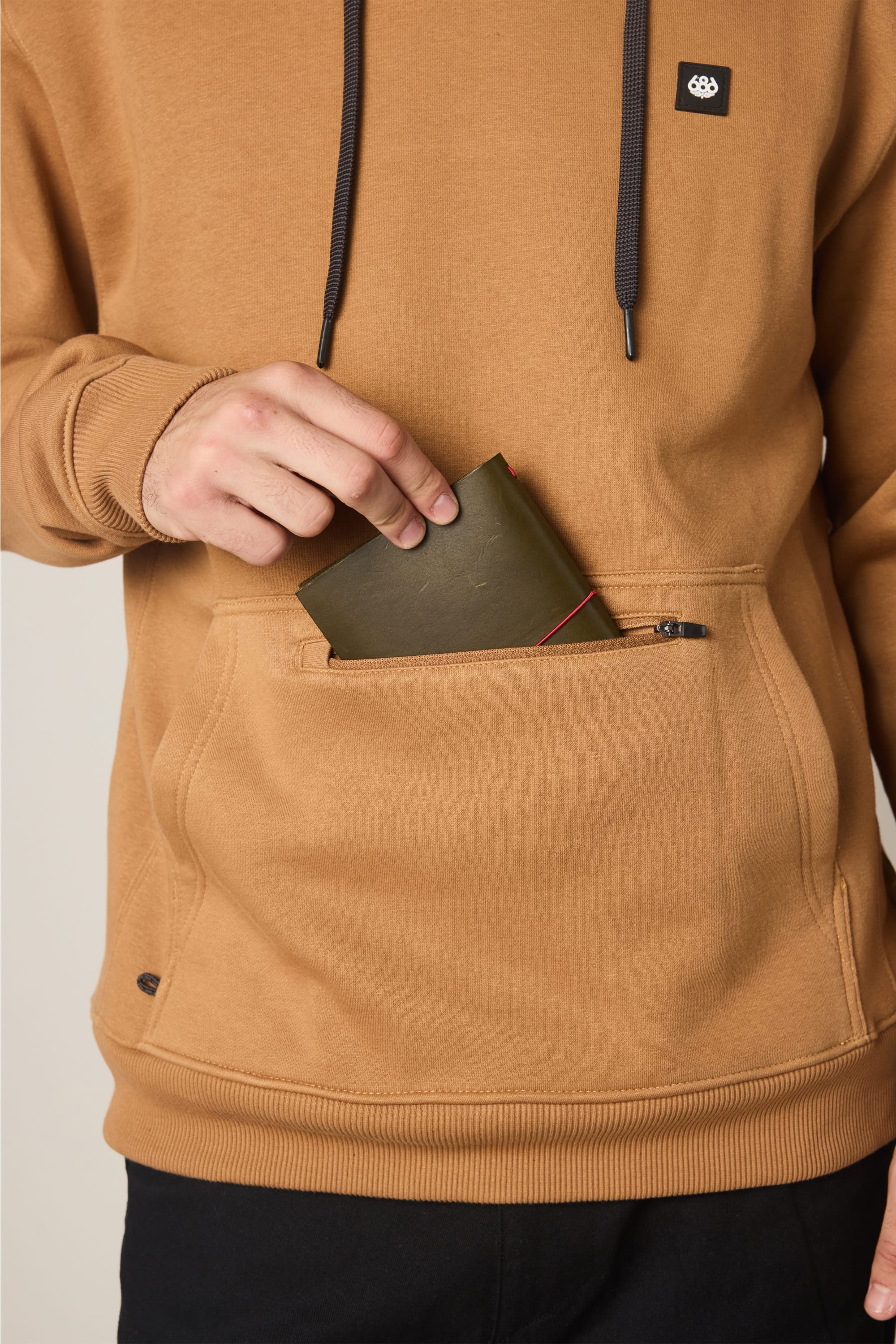 A person wearing the 686 Men's All Season Hoody in tan places an olive green wallet into the front pouch pocket.