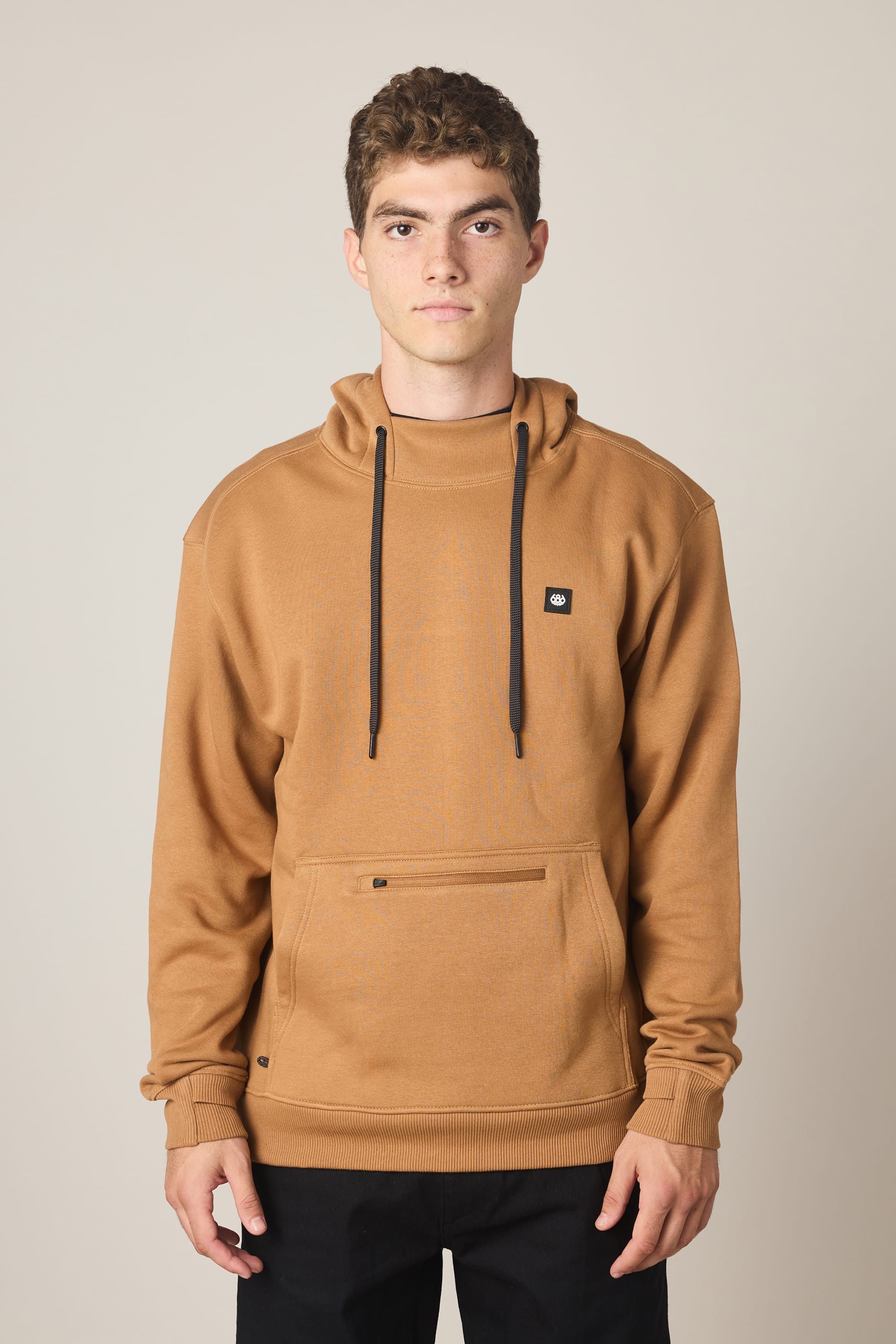 A young man with short curly hair wears the 686 Men's All Season Hoody—a tan sweatshirt with black drawstrings and a front pocket—standing against a plain light background, looking directly at the camera with a neutral expression.