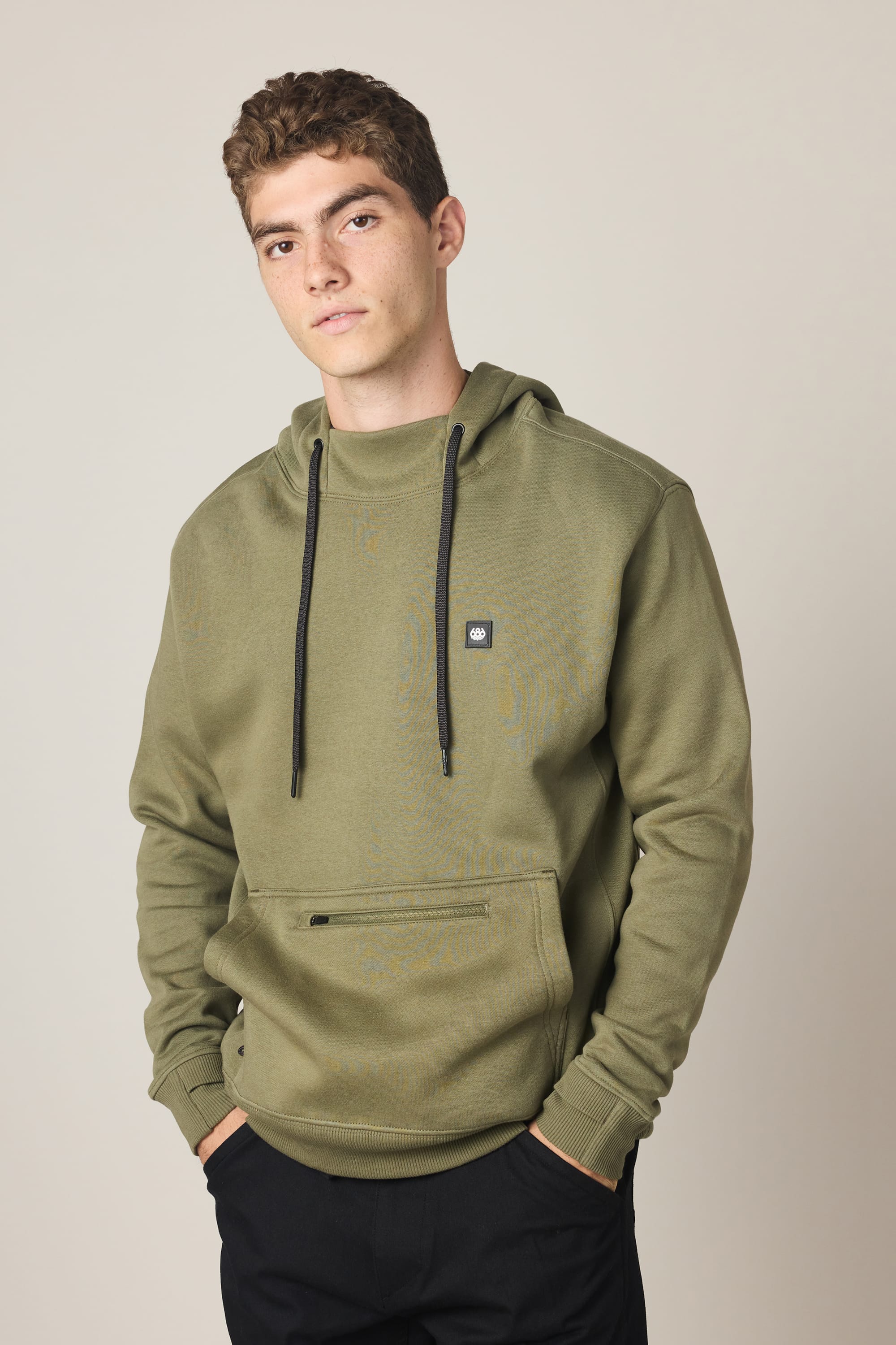 A young man with short curly hair wears the 686 Men's All Season Hoody in olive green with black drawstrings and a small chest logo, paired with black pants, standing hands-in-pocket against a plain light background.