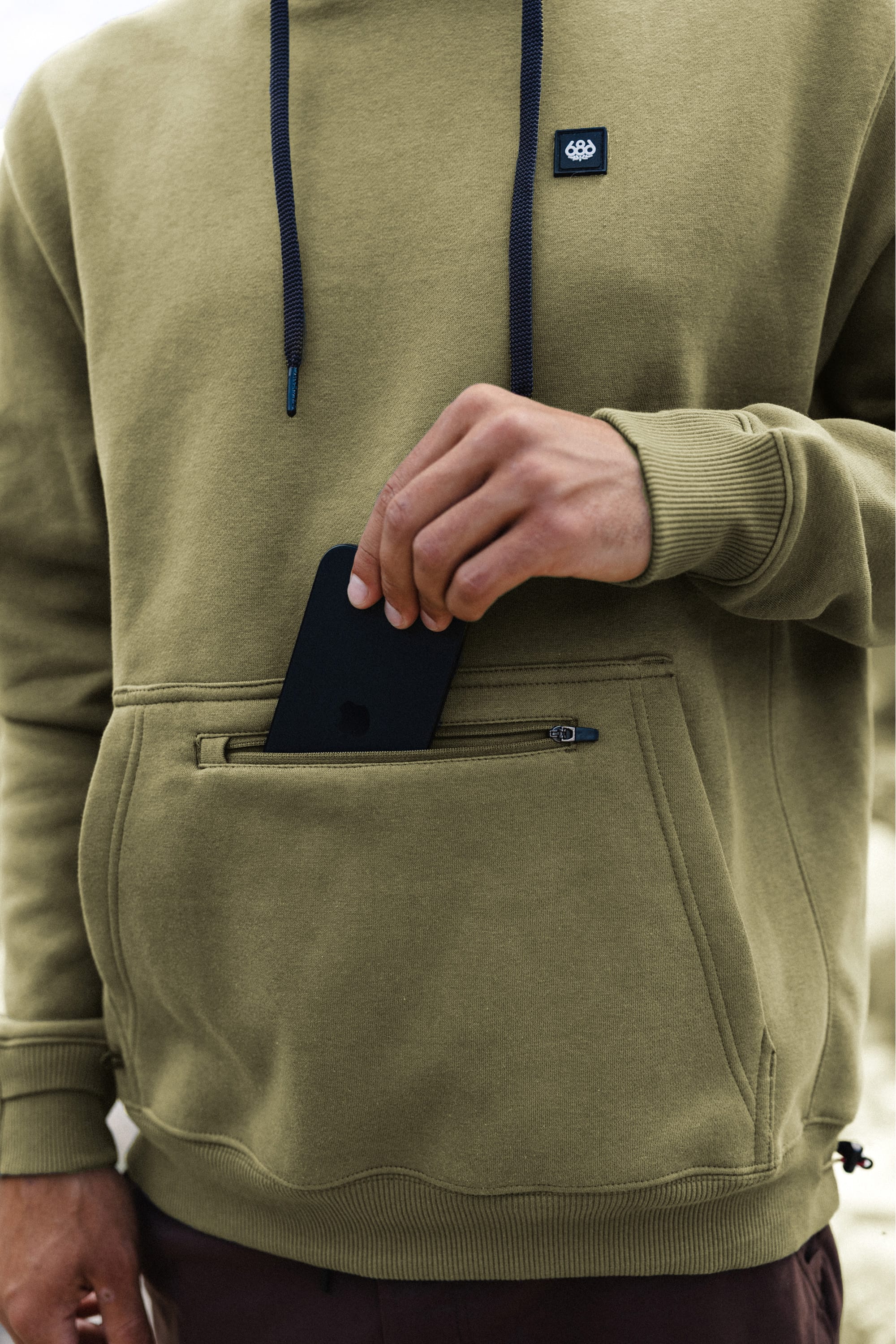 Wearing the 686 Men's All Season Hoody in olive green, a person places a black smartphone into the zippered front pocket of the hoodie. The photo centers on their torso and hand, with their face out of view.