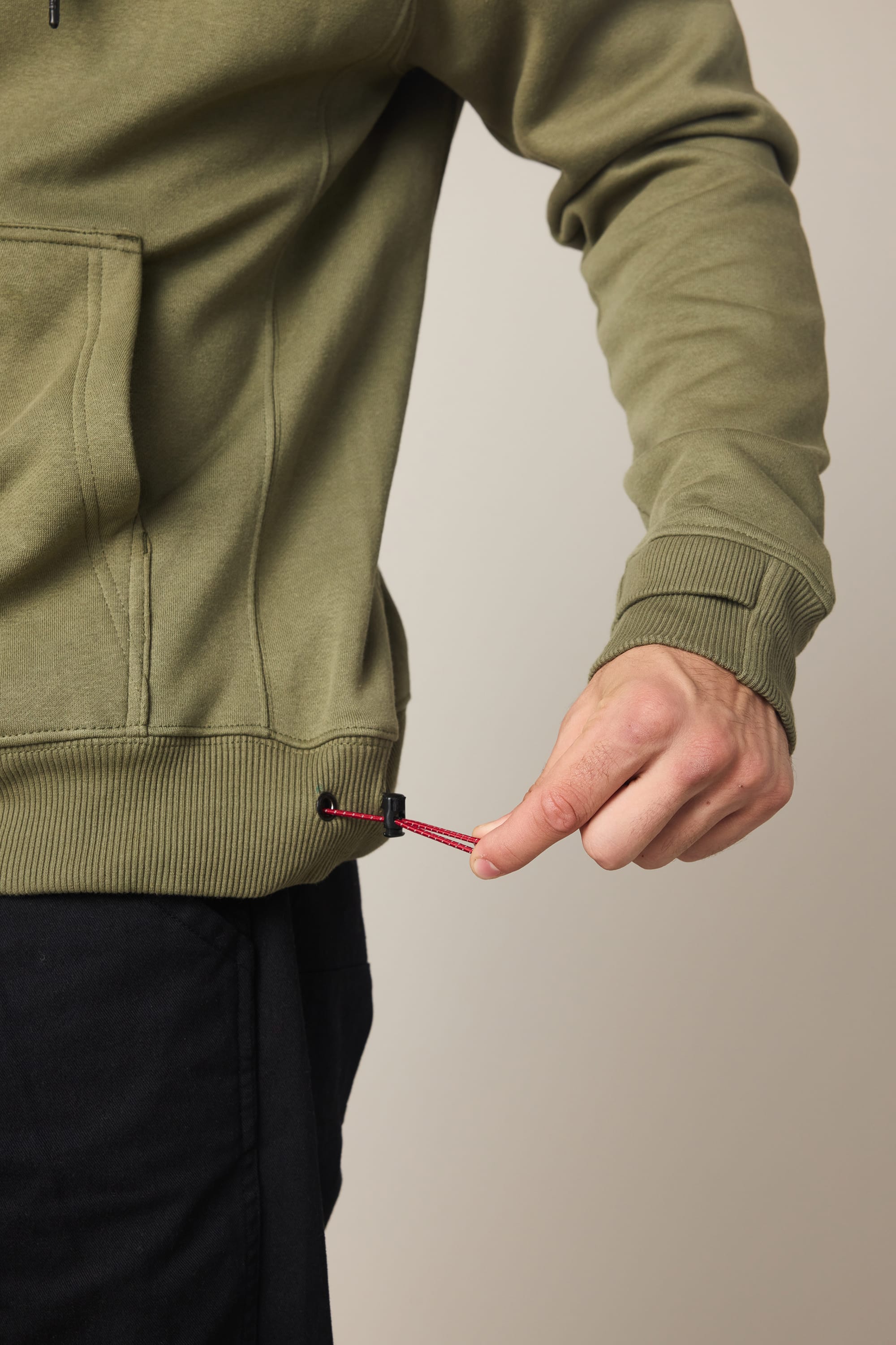 A person adjusts the red drawstring on the hem of a 686 Men's All Season Hoody in olive green, paired with black pants. Only their torso and hand are visible against a neutral background.