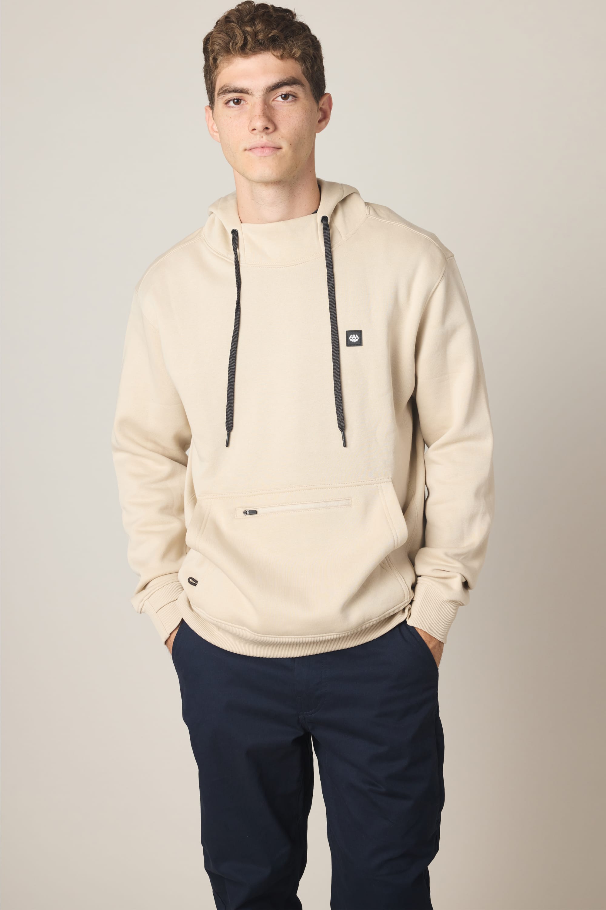 A young man with short curly hair wears the 686 Men's All Season Hoody in beige with black drawstrings and a small logo patch, paired with dark navy pants, standing hands-in-pockets against a plain background.