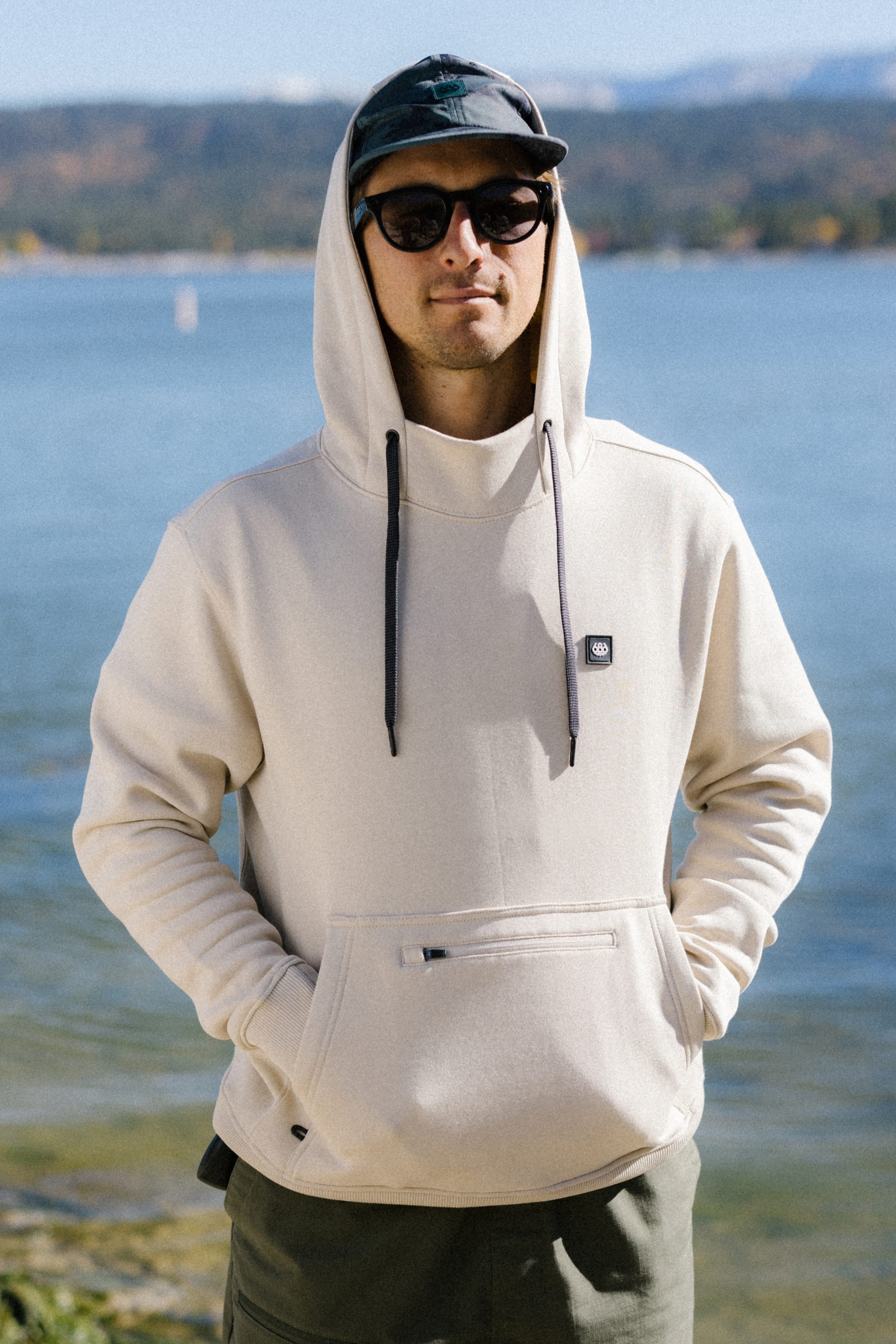 A person wearing sunglasses, a cap, and the 686 Men's All Season Hoody by 686—crafted from soft cotton and spandex—stands outdoors near a lake with mountains and trees in the background on a sunny day.