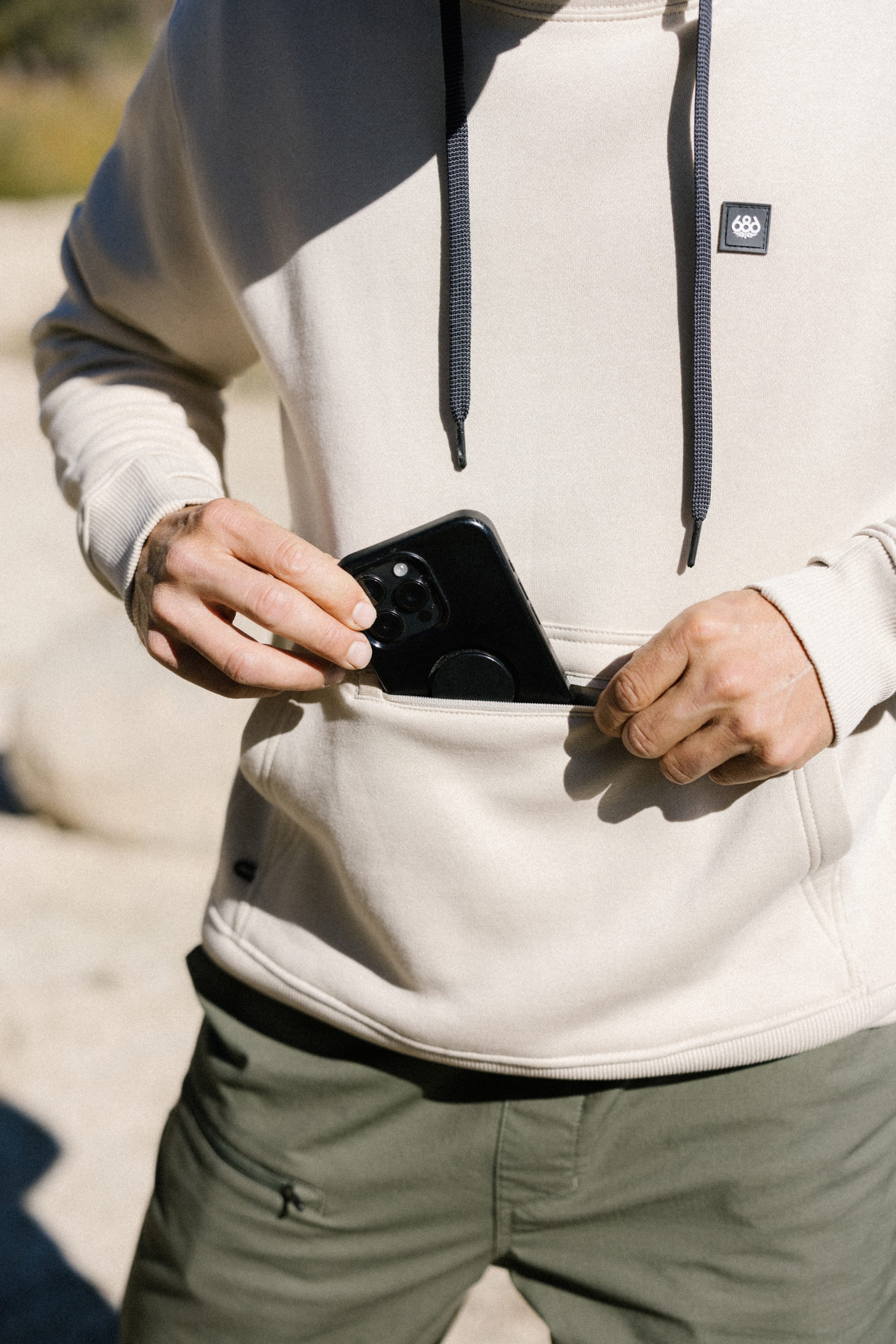 A person wearing a 686 Men's All Season Hoody in beige and green pants places a black smartphone into the front pocket of the hoody outdoors.