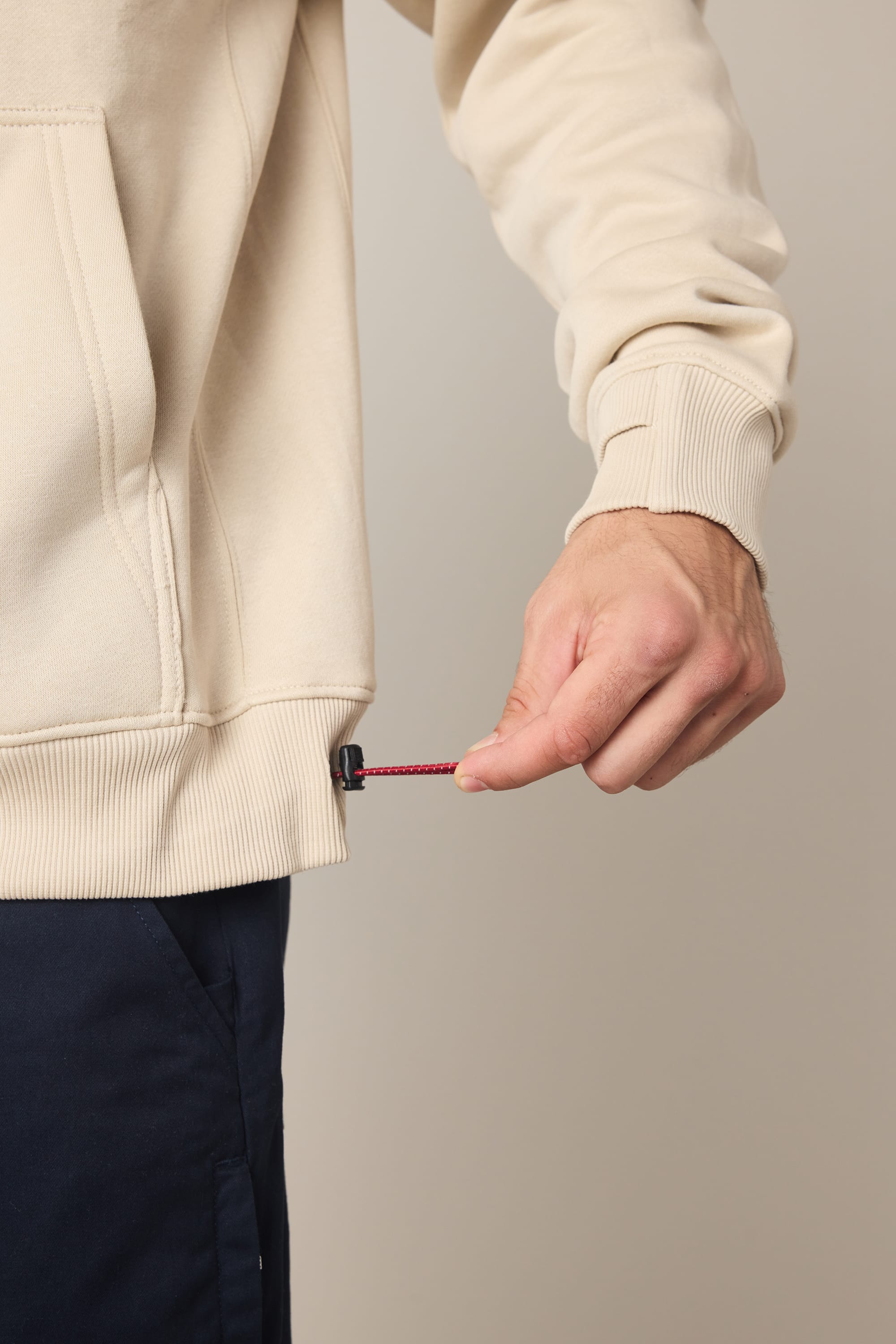 A person wearing the 686 Men's All Season Hoody in beige adjusts the red drawstring at the hem with their right hand.