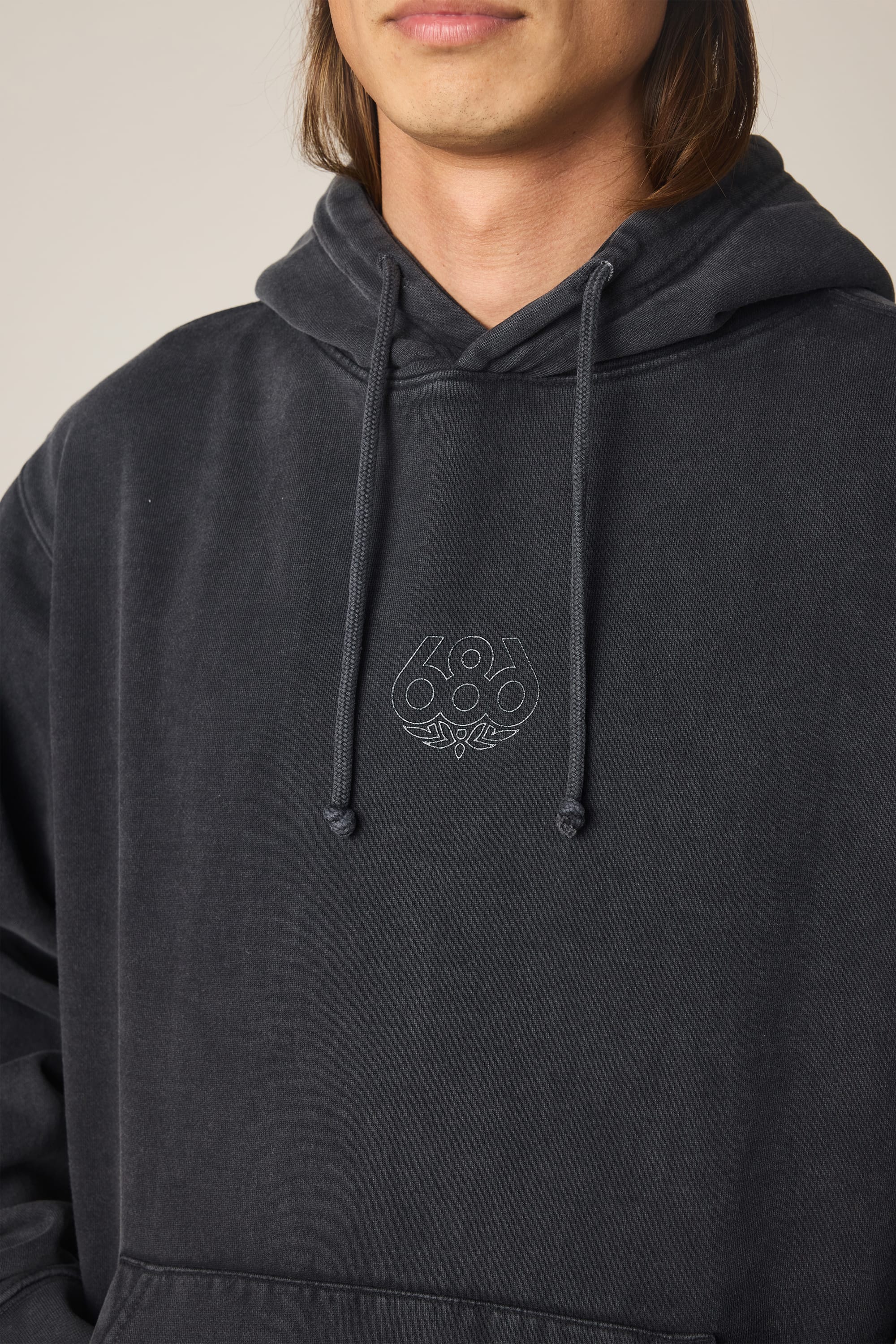 A person wearing the 686 Men's High Def Relaxed LS Hoody in dark gray, featuring a small 686 logo and script text embroidered on the chest; only their lower face and upper torso are visible.