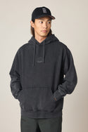 A person wearing a black baseball cap and the 686 Men's High Def Relaxed LS Hoody, featuring a small embroidered logo, stands with hands in the front pocket, looking to the side against a plain light background.