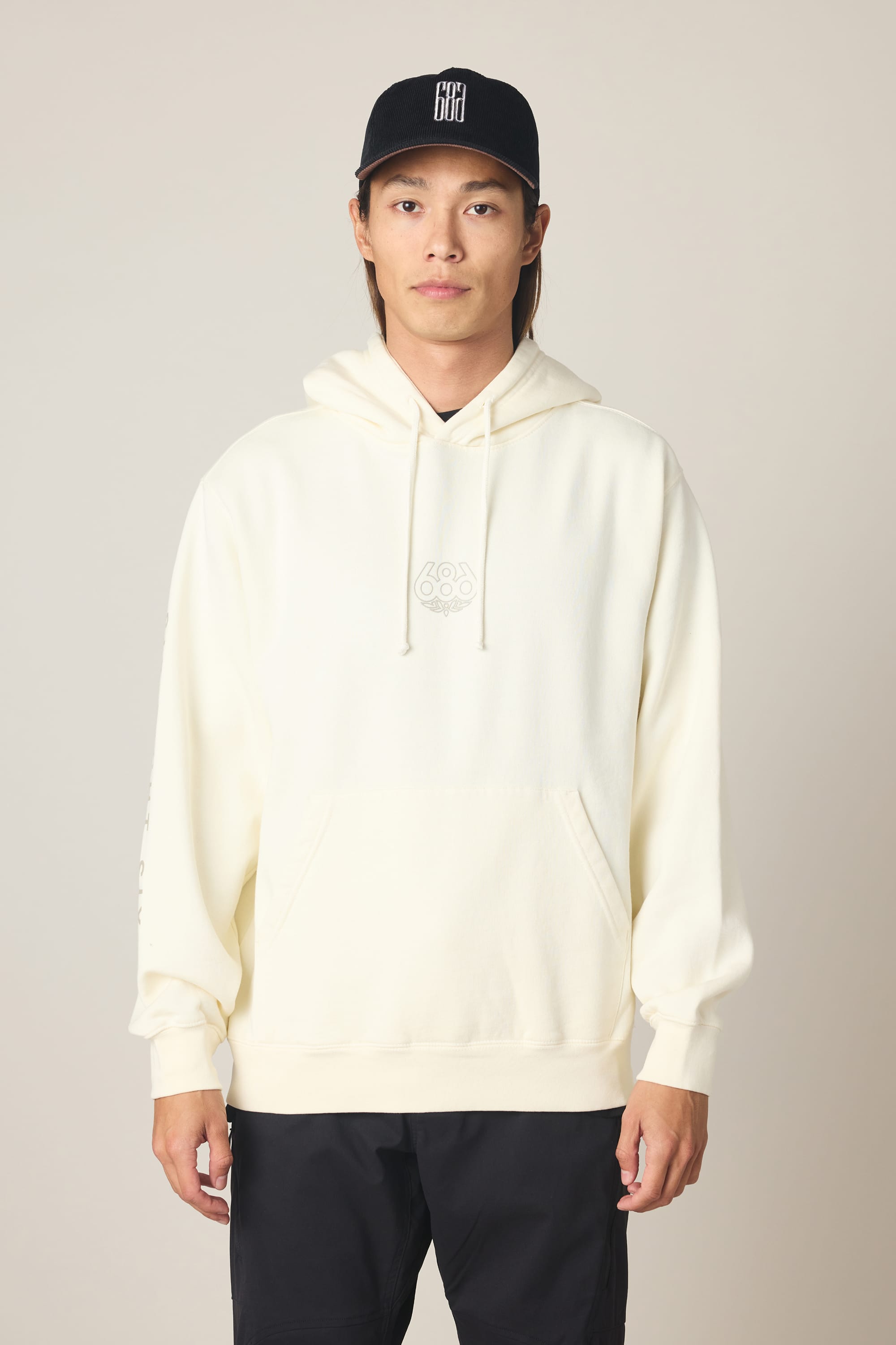 A person wearing the 686 Men's High Def Relaxed LS Hoody in cream and a black baseball cap stands facing forward against a neutral background.