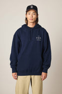 A person stands facing forward in a 686 Men's Unwork Spec Athletic LS Hoody (navy blue) with a small white logo and text on the left chest, beige pants, and a black baseball cap with a white design. The background is plain and light-colored.
