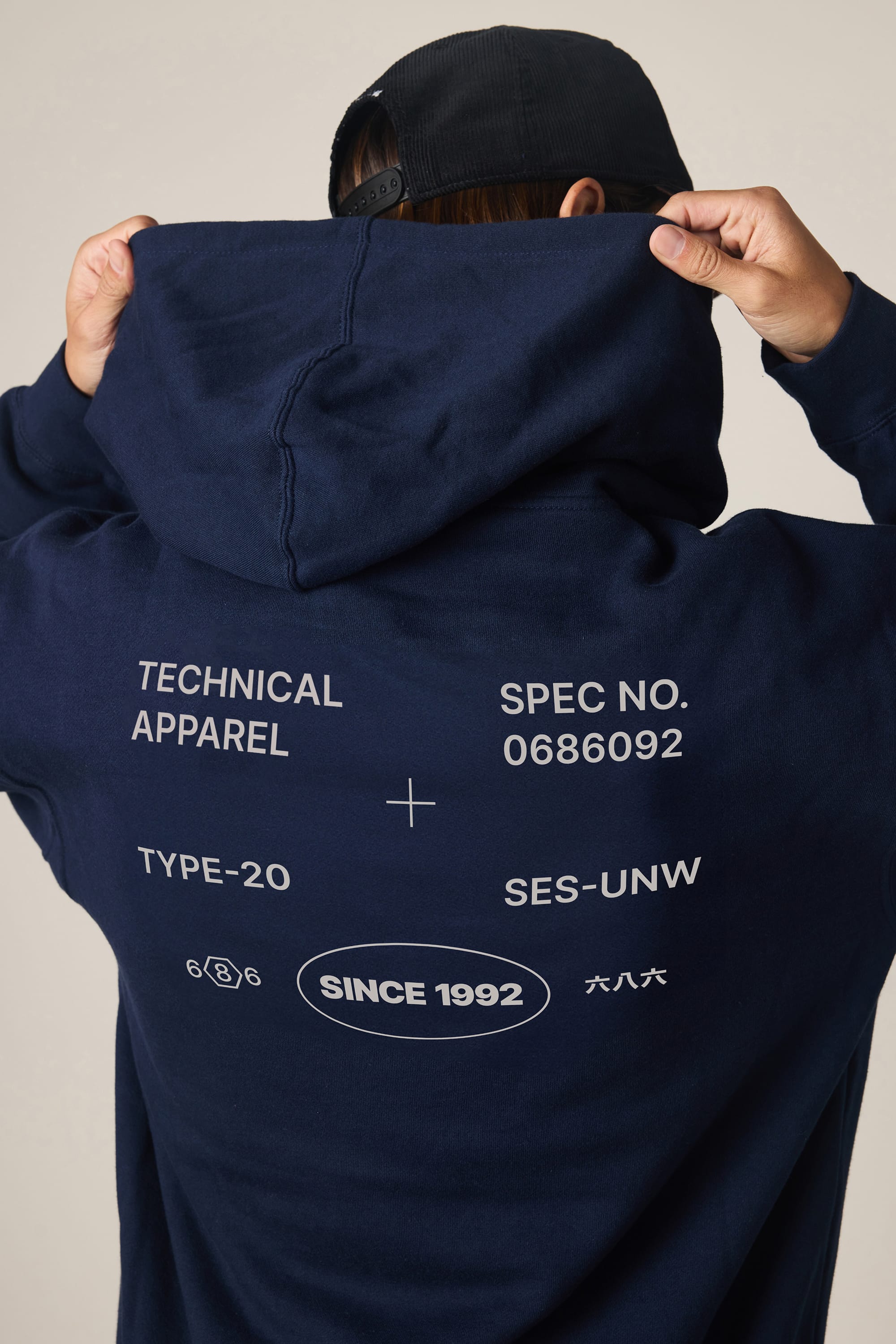 A man wearing the 686 Men's Unwork Spec Athletic LS Hoody in navy holds up the hood. The back of this 686 sweatshirt features white printed text: TECHNICAL APPAREL, SPEC NO. 0686092, TYPE-20, SES-UNW, and SINCE 1992 along with smaller symbols. He also wears a black cap.