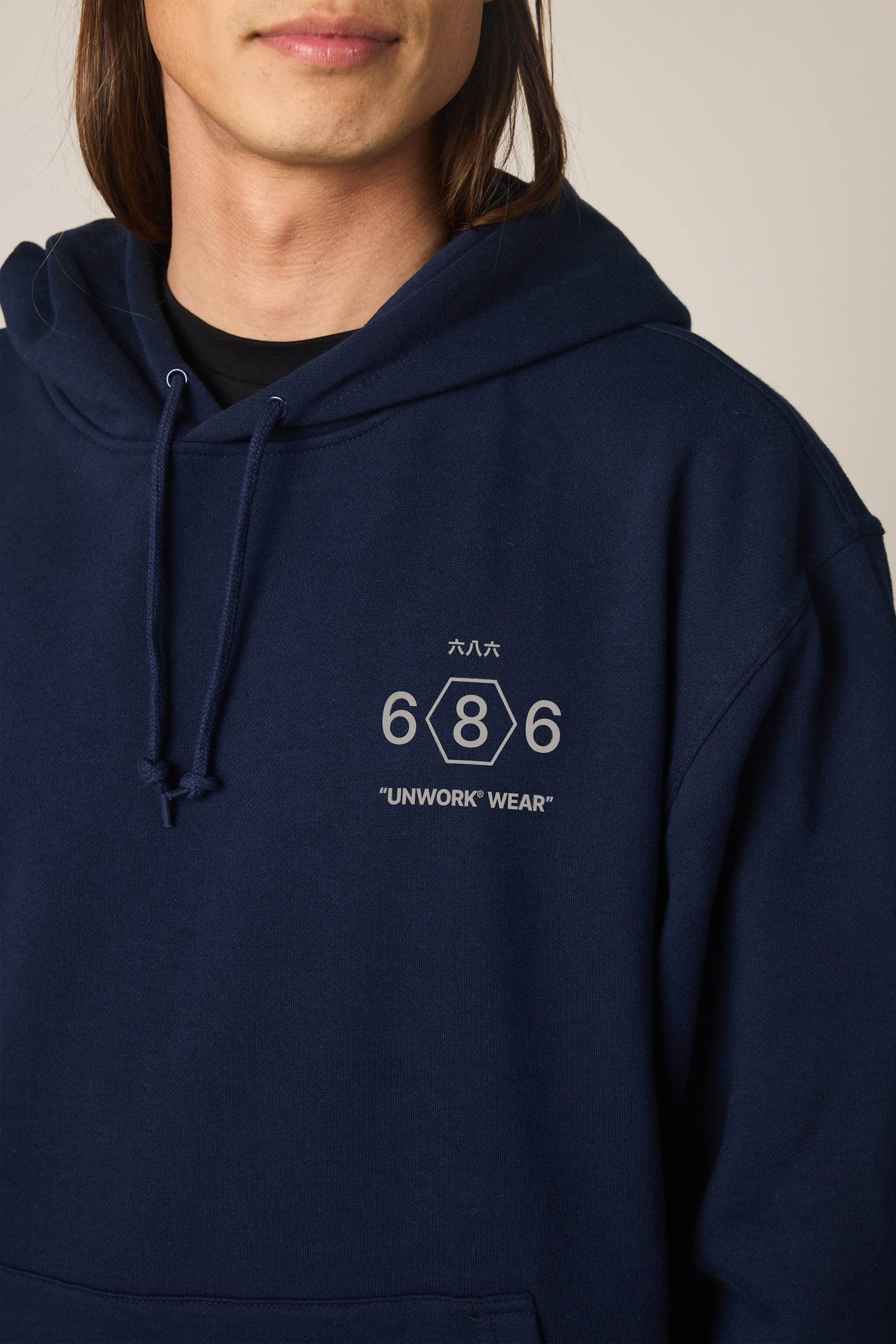 A person wearing the 686 Men's Unwork Spec Athletic LS Hoody in navy blue, featuring the 686 brand name, "UNWORK WEAR" text, Japanese characters, and a hexagon design printed on the chest. Only their lower face and upper torso are visible.